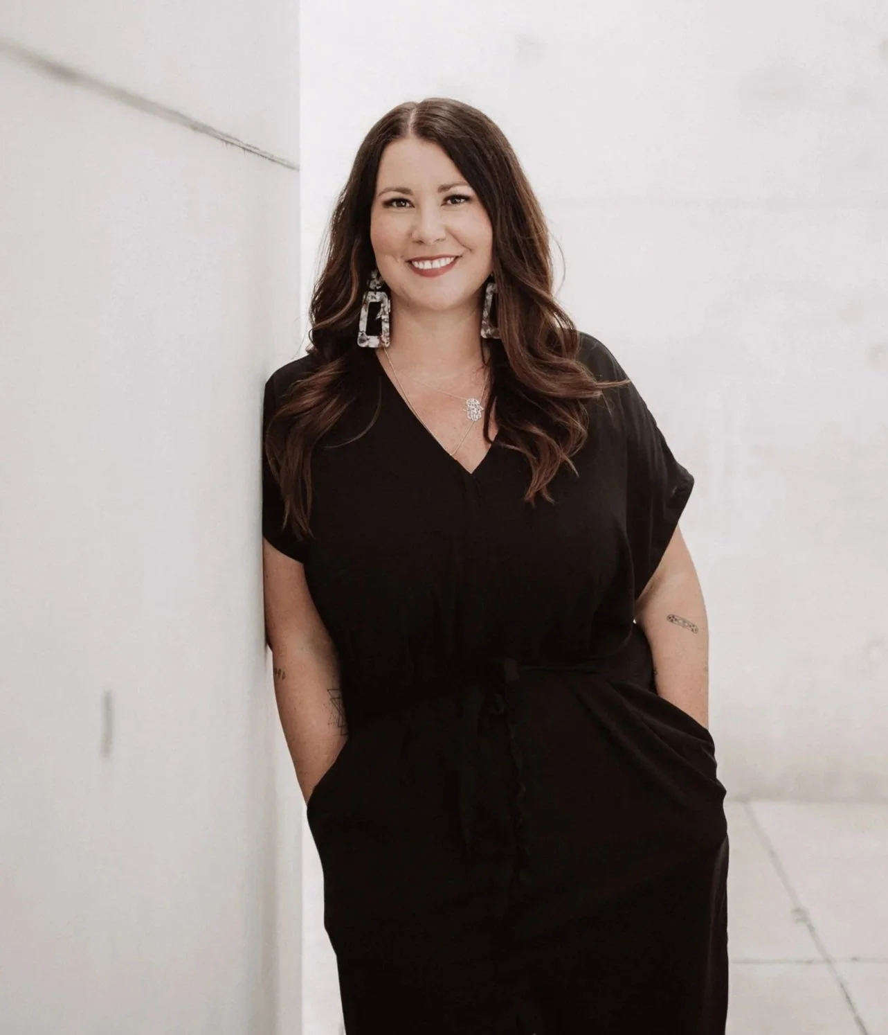 Mirja Marek. Eine lächelnde Frau mit braunen Haaren in einem schwarzen Kleid lehnt an einer weißen Wand in einem hellen Raum.