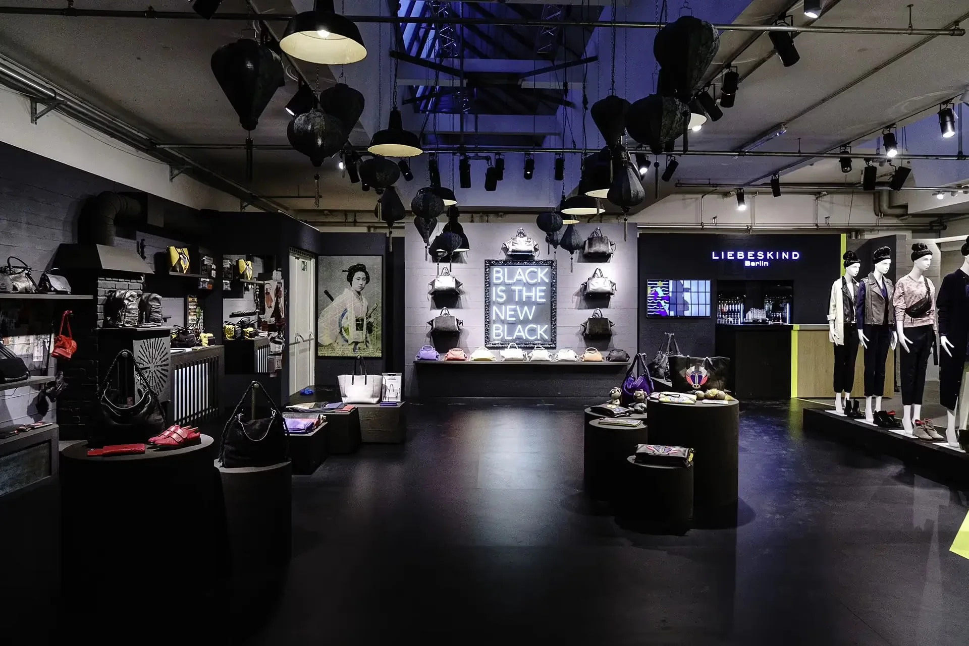 Ein Modegeschäft mit schwarzen, weißen und leuchtenden Akzentfarben. Es sind Taschen, Handtaschen und Kleidung ausgestellt, mannequin tragen Kleidungsstücke. Die Wand hat ein Neon-Schild mit der Aufschrift "BLACK IS THE NEW BLACK".