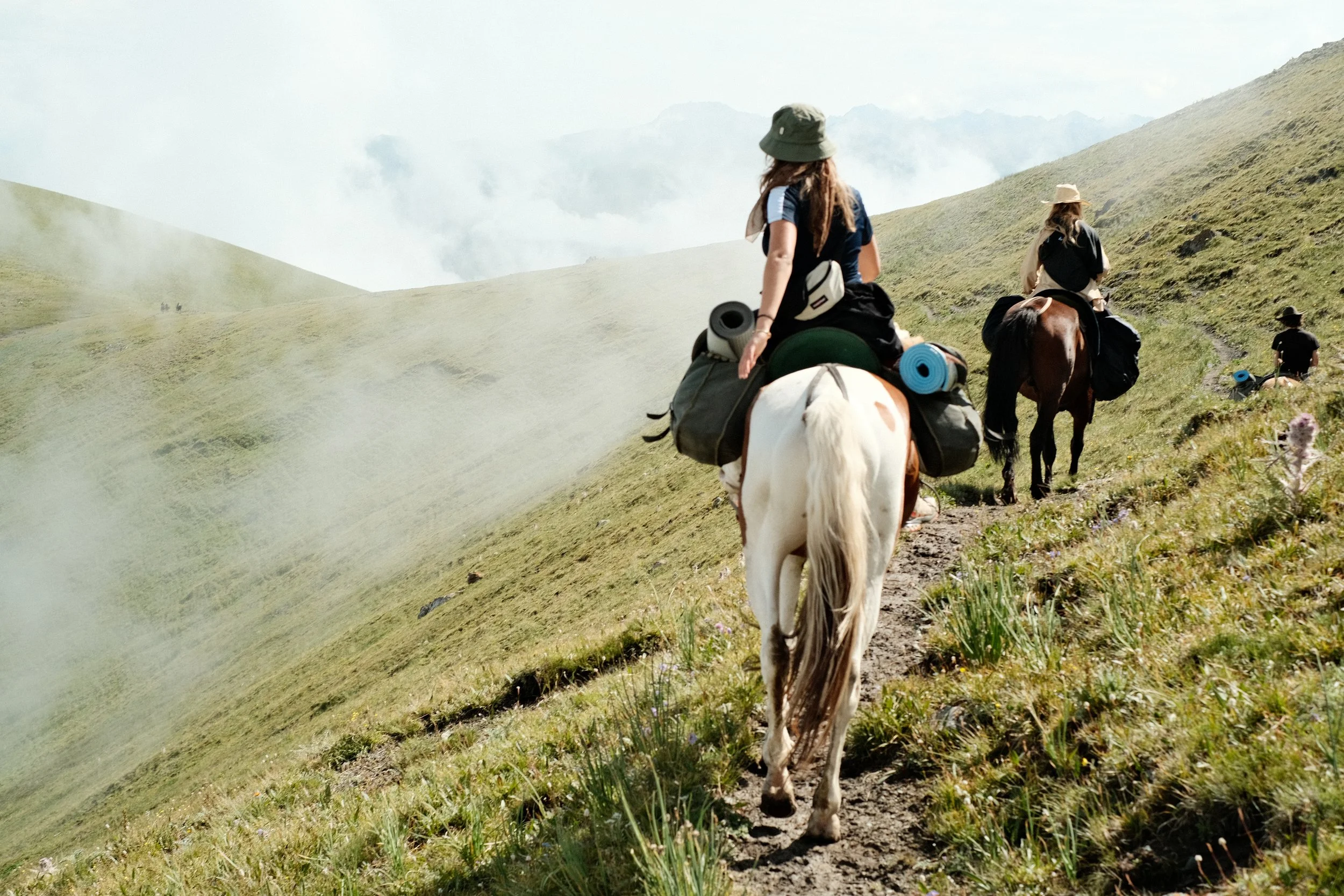 People riding horses along a mountain trail through grassy hills with mist and clouds in the background.