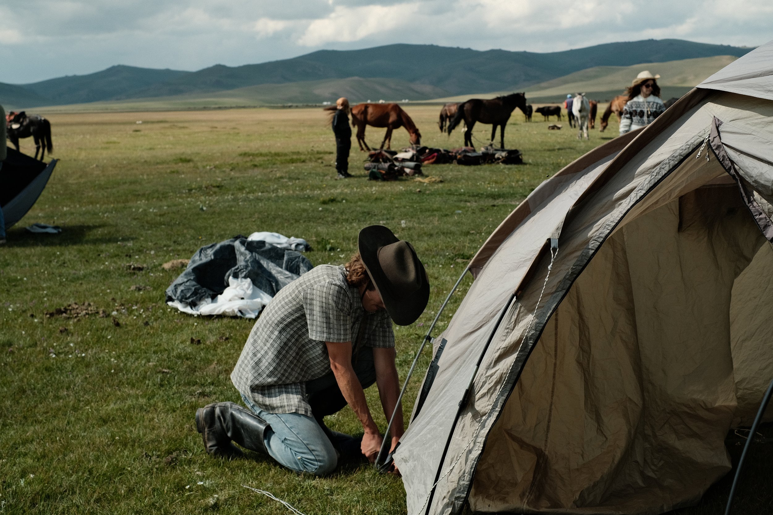 A person kneeling on the grass, preparing a tent, with horses and other campers in the background in a wide open field with mountains in the distance.
