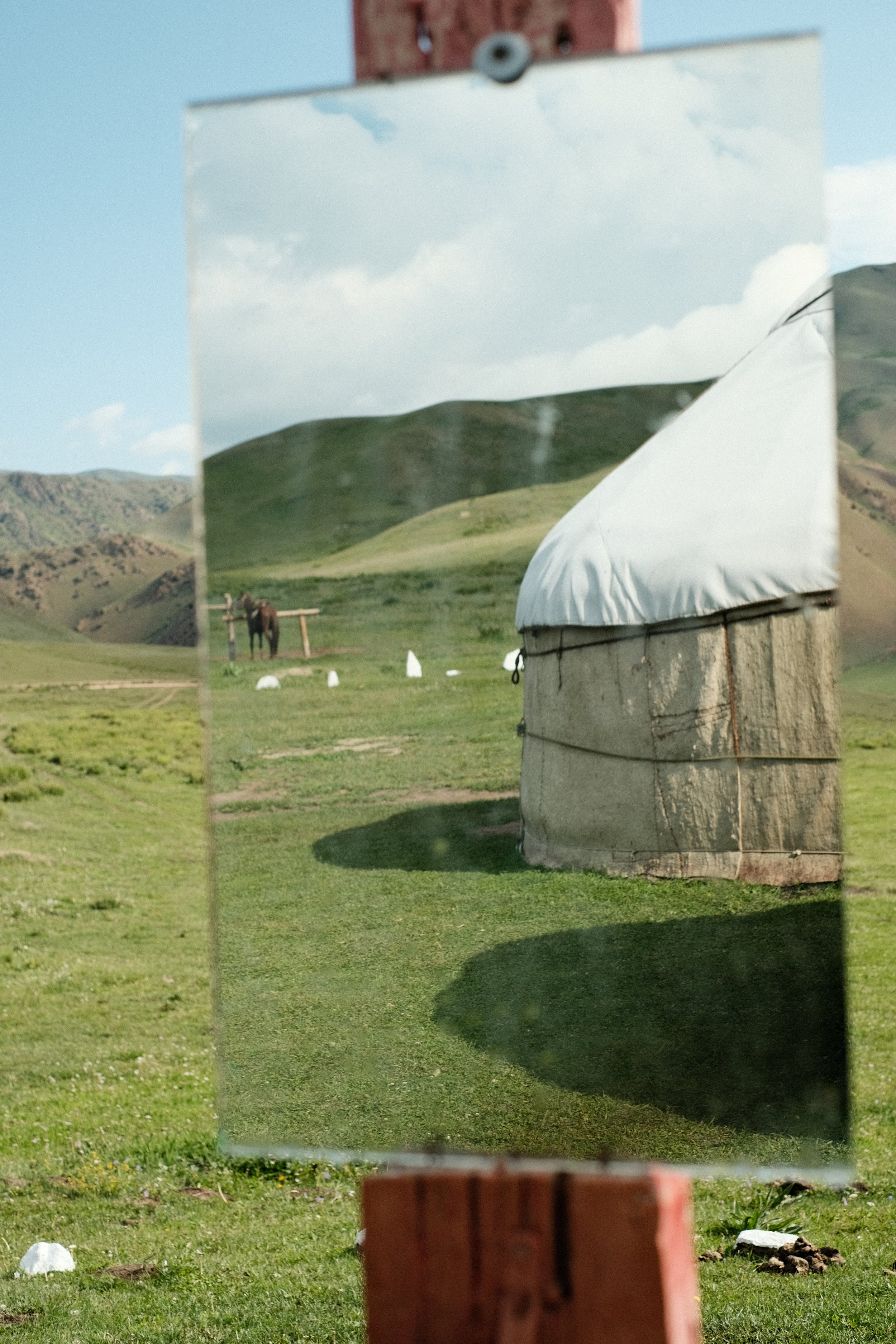 A mirror reflecting a landscape of green hills, a horse, and a white yurt, with the mirror mounted on a red brick structure in an open field.