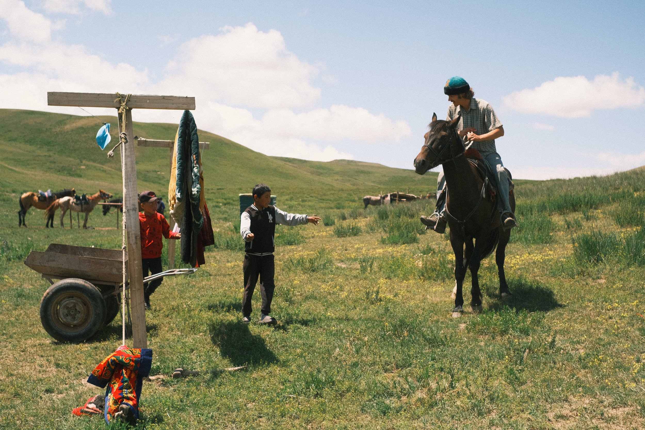 Two children and a man on horseback in a grassy field with rolling hills, a makeshift clothesline, and additional horses in the background under a partly cloudy sky.