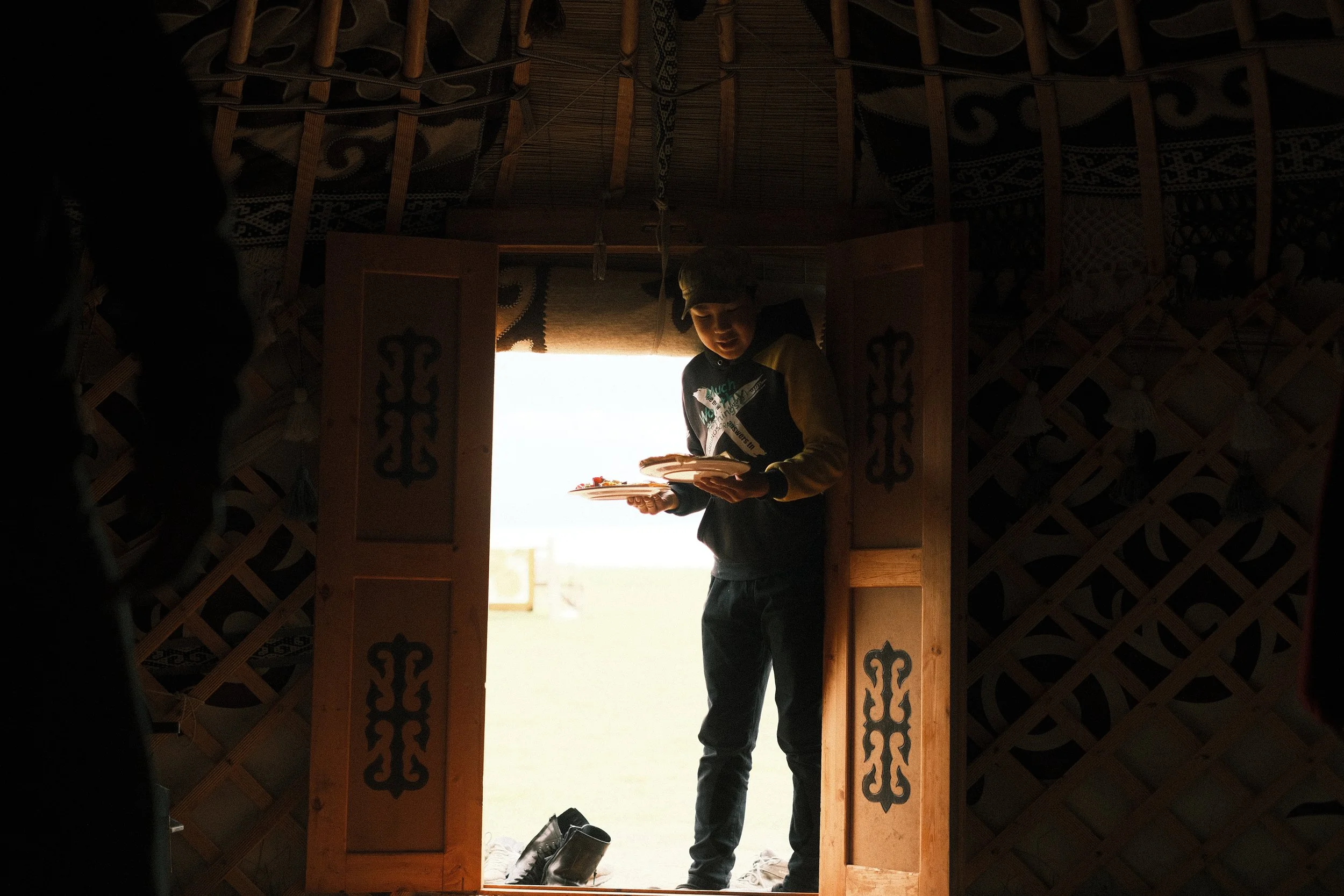 A young man standing in the doorway of a traditional yurt, holding a plate of food. The interior of the yurt has wooden lattice walls and decorative panels on the door. Shoes are placed outside the entrance, and sunlight is streaming in from outside.