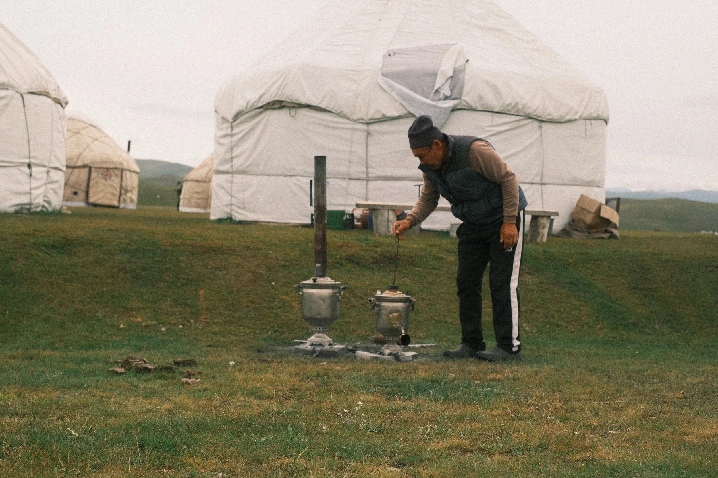 A man in a beanie and jacket tending to two small traditional outdoor stoves on grass, with large dome-shaped yurts in the background