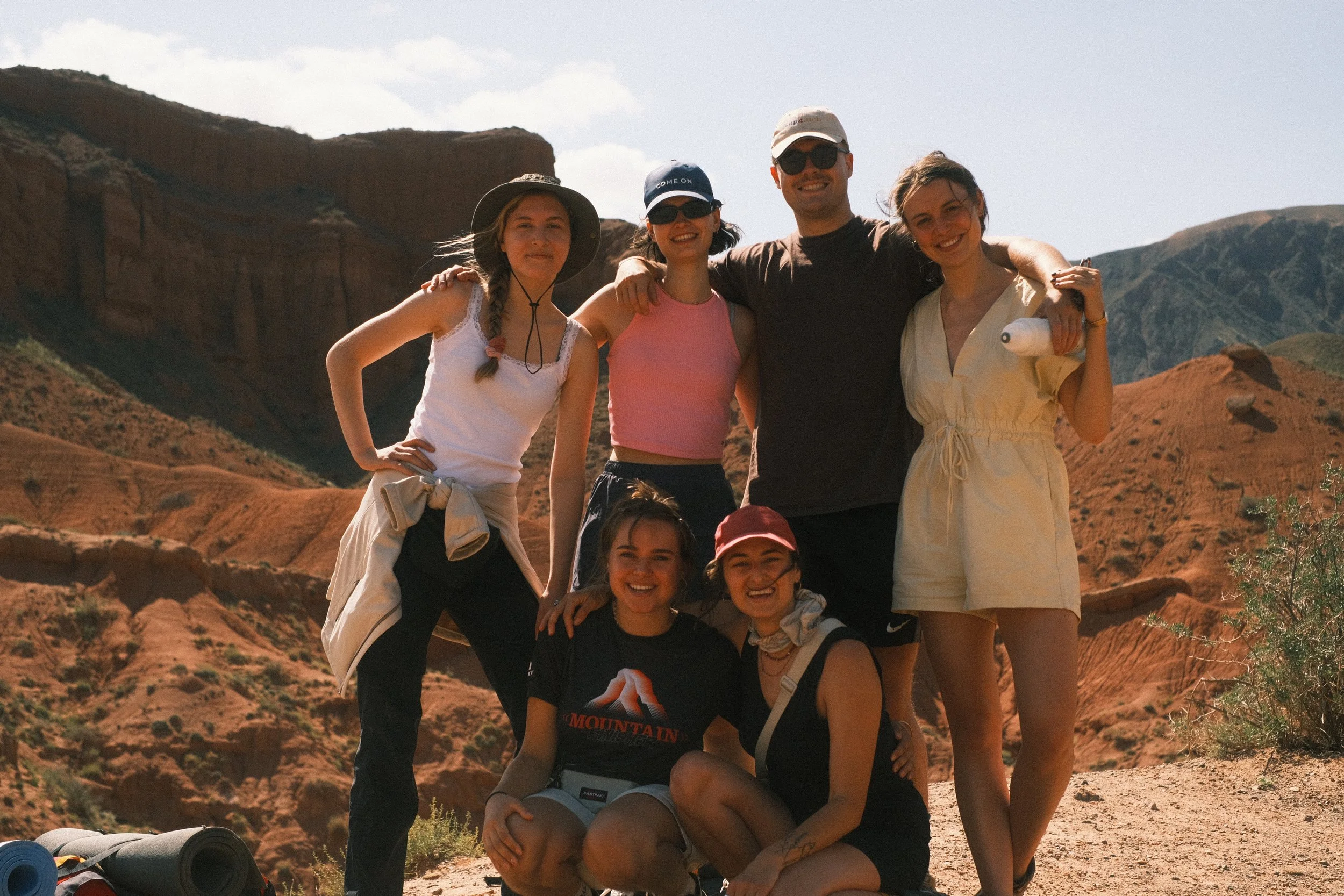 Group of six friends hiking in a desert landscape with red rock formations in the background.