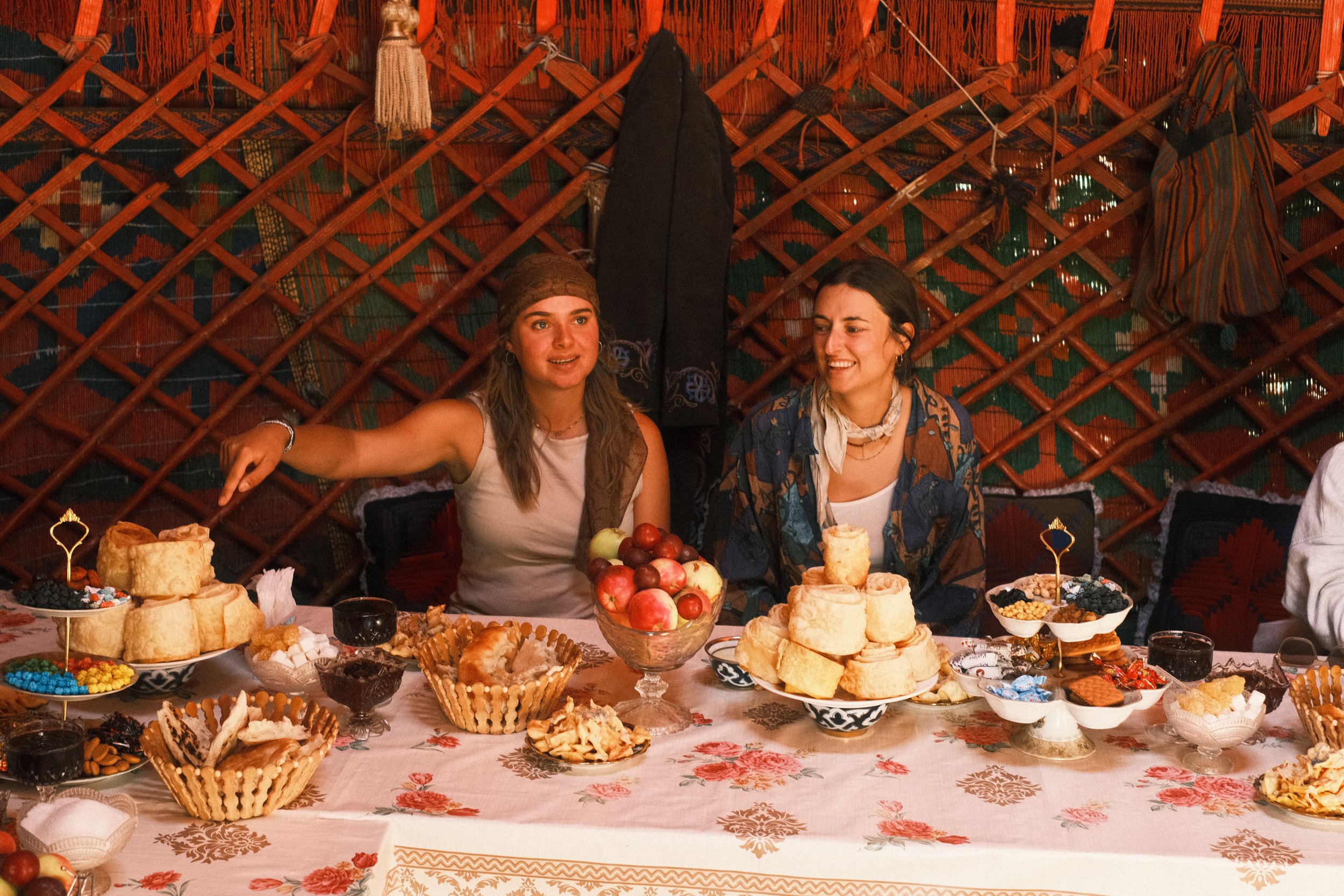 Two women sitting at a table filled with baked goods, fruit, and desserts, inside a decorated yurt, engaging in conversation and smiling.
