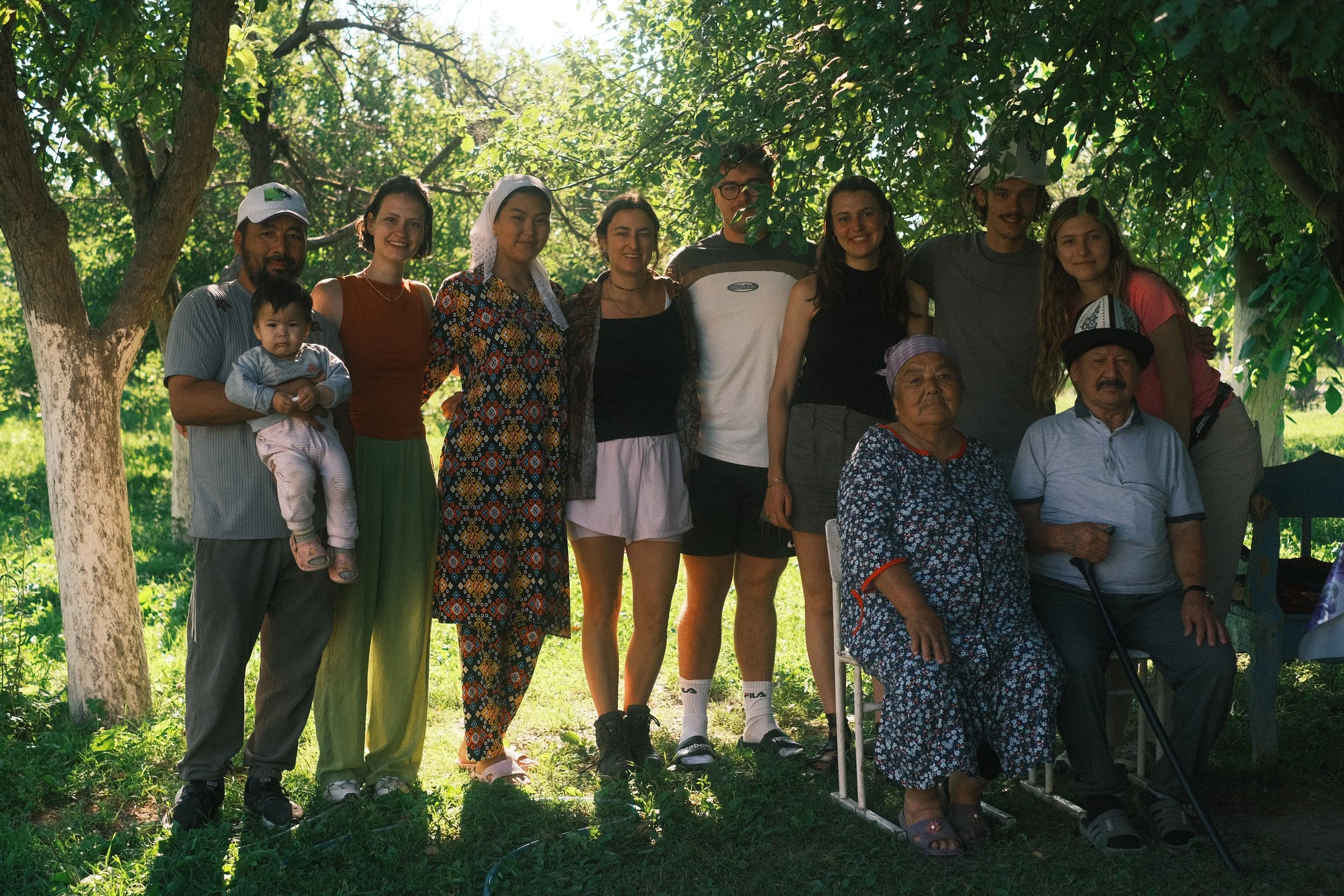 A diverse group of people posing outdoors in a shaded area with trees, including adults, teenagers, and elderly individuals, some sitting and some standing.
