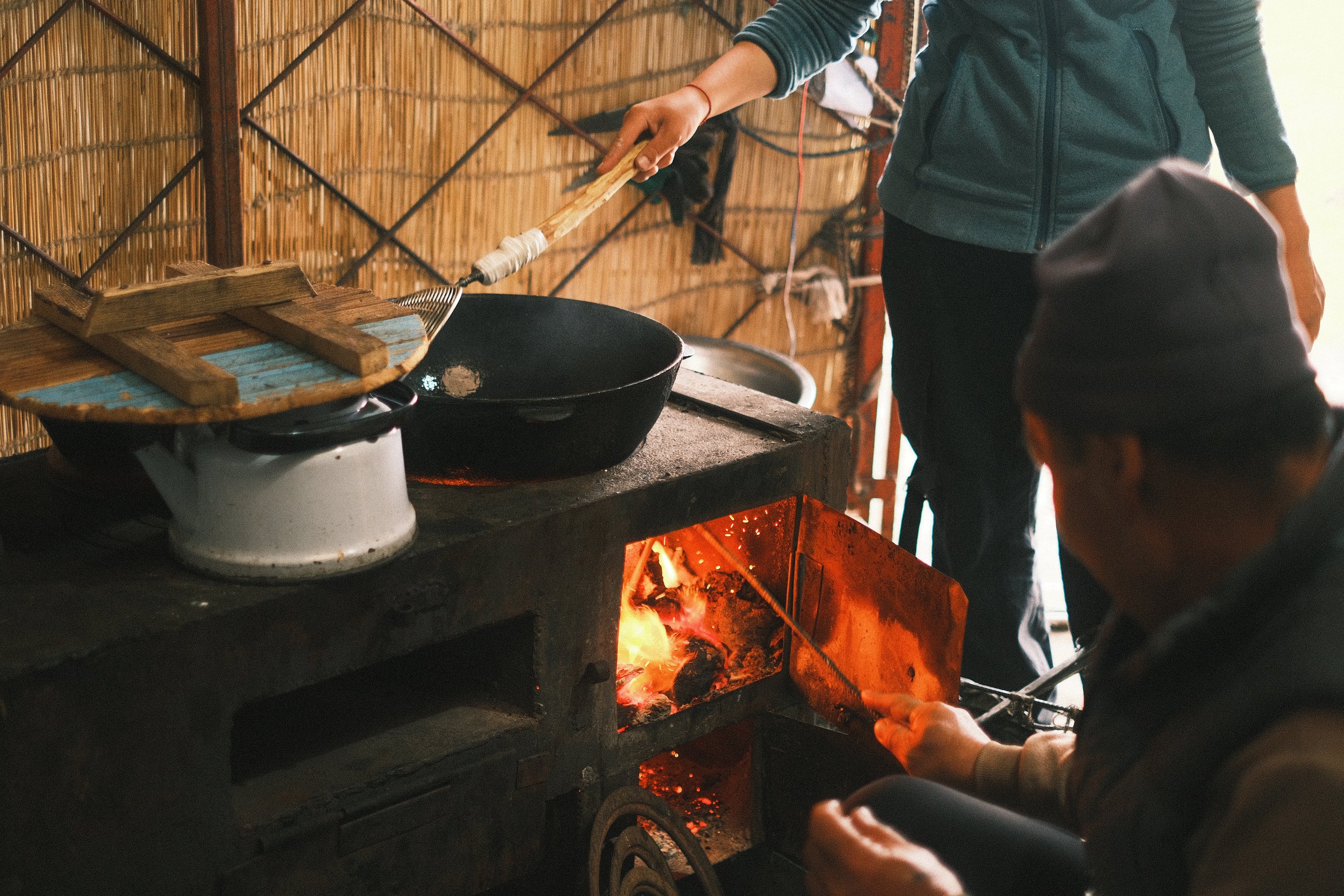 Two people cook over an open fire in a rustic kitchen with bamboo walls; one stirs a black cast iron pan, and the other adjusts a heat source with a tool.