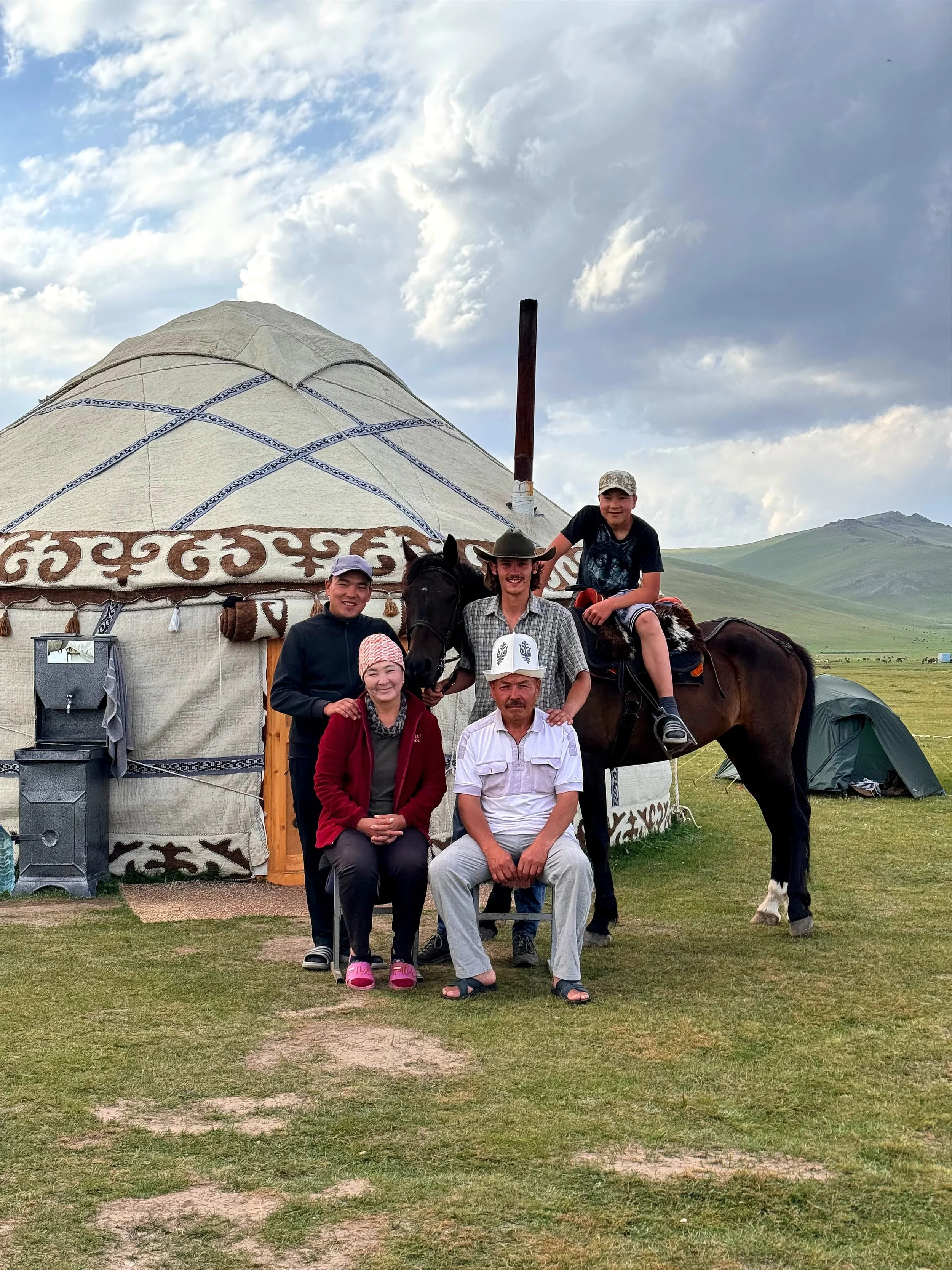 A group of five people and a horse in front of a traditional yurt in a grassy field with mountains and cloudy sky in the background.