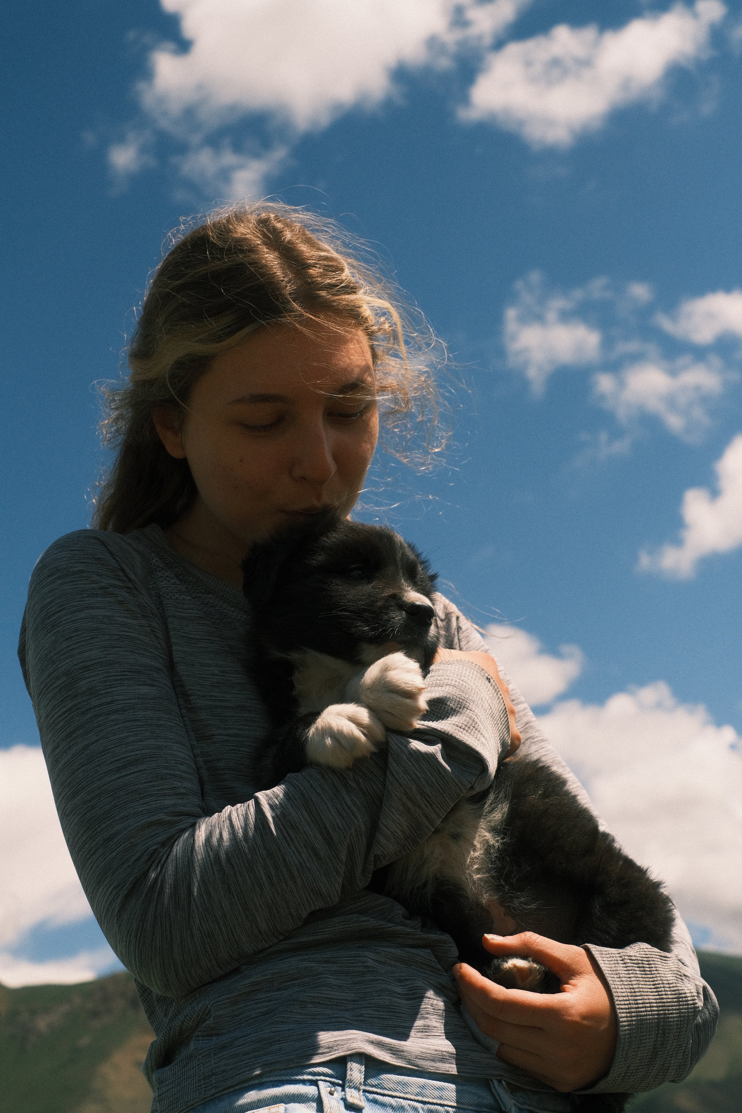 A woman holding a black and white puppy outdoors under a blue sky with clouds.