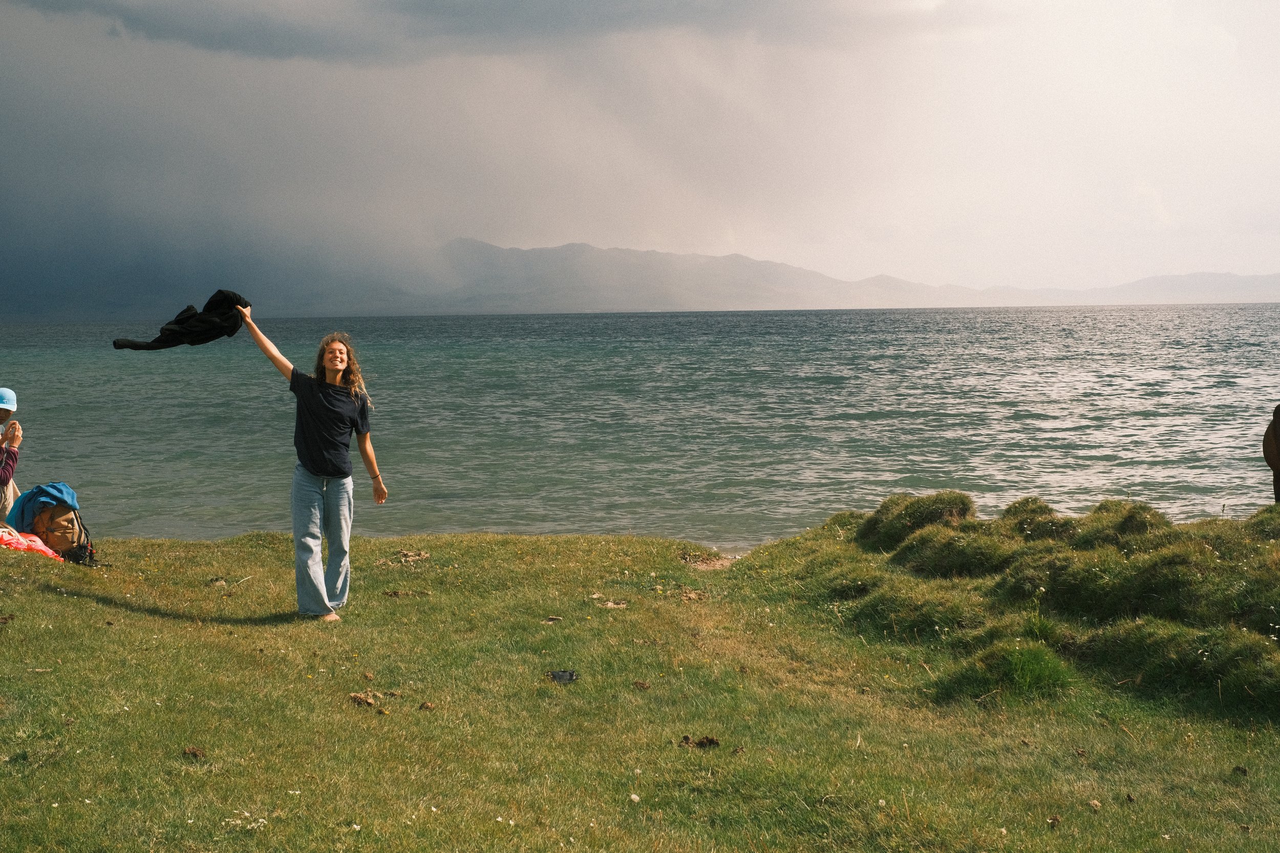 A woman standing on a grassy area near a large body of water, smiling and waving a black piece of clothing in the air, with a cloudy sky and distant mountains in the background.
