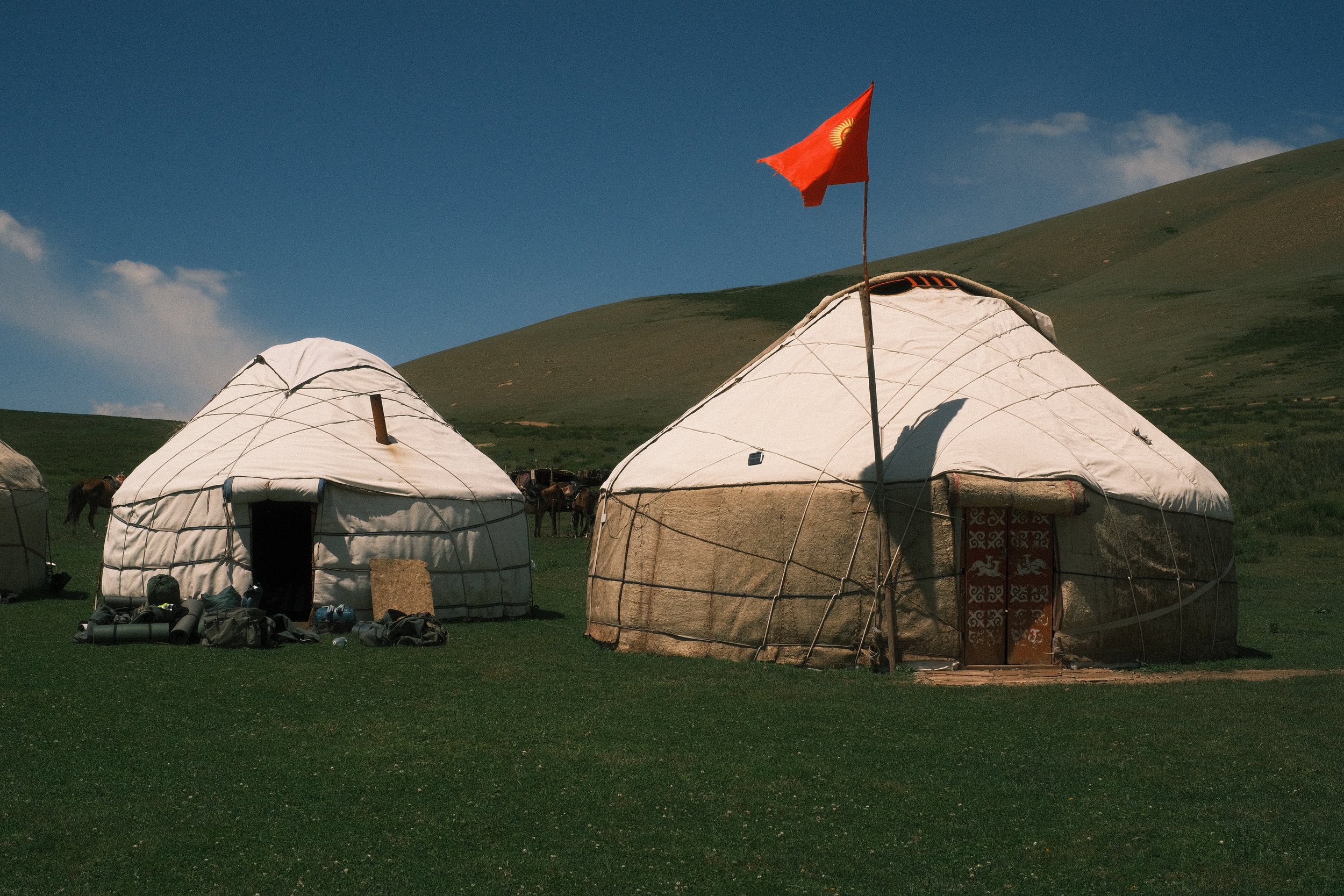 Two traditional yurts set up on grassy land with a mountain in the background. One yurt has a flag on a pole, and there are backpacks and camping gear in front of the yurts.
