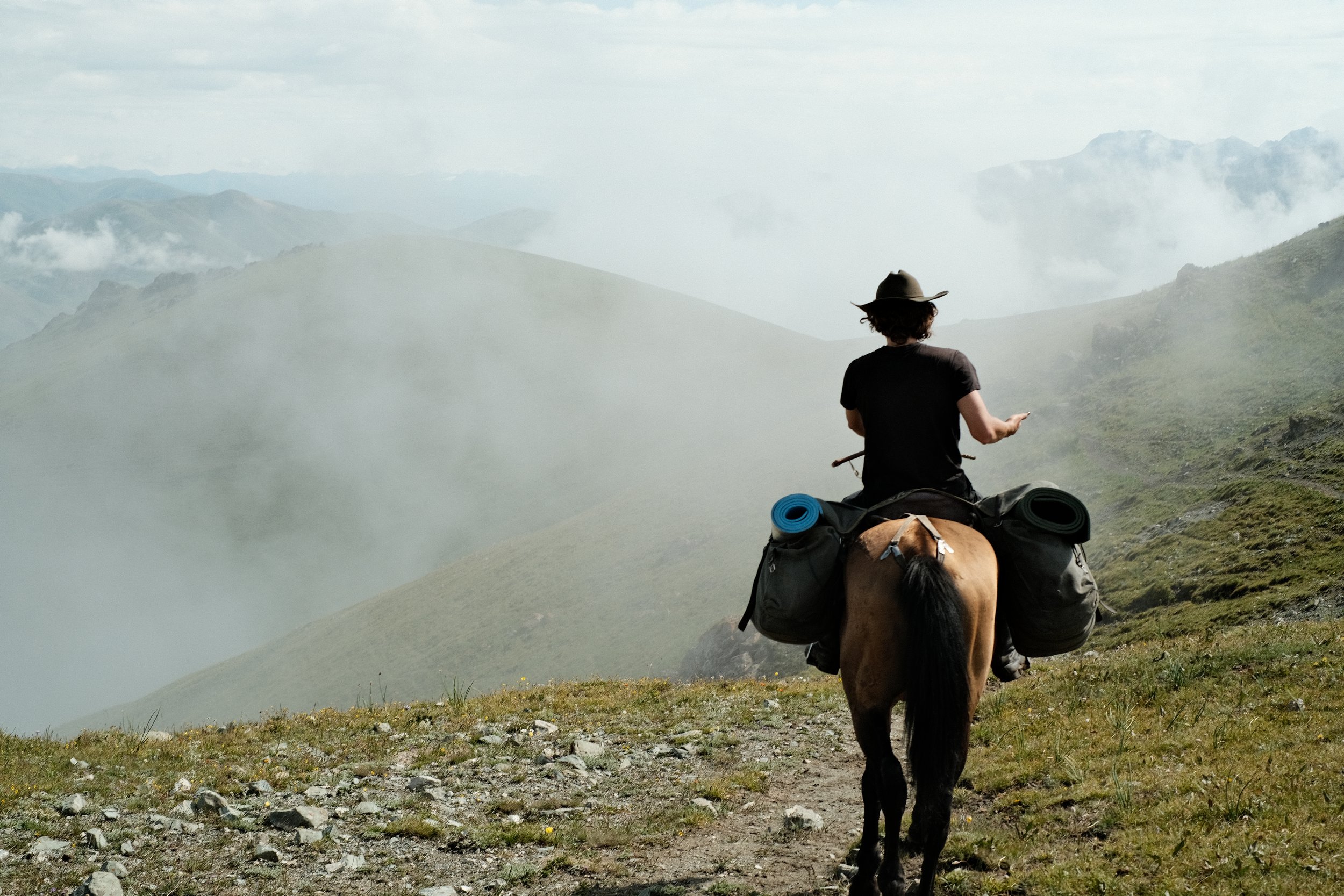 Person riding a horse on a mountain trail during cloudy weather, with mountains and clouds in the background.