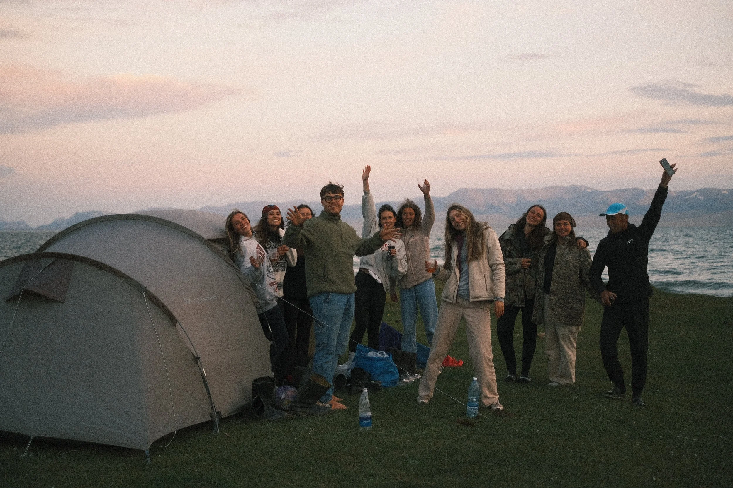 Group of friends enjoying a camping trip by a lake at sunset, standing near tents, with mountains in the background, some holding drinks, and smiling at the camera.