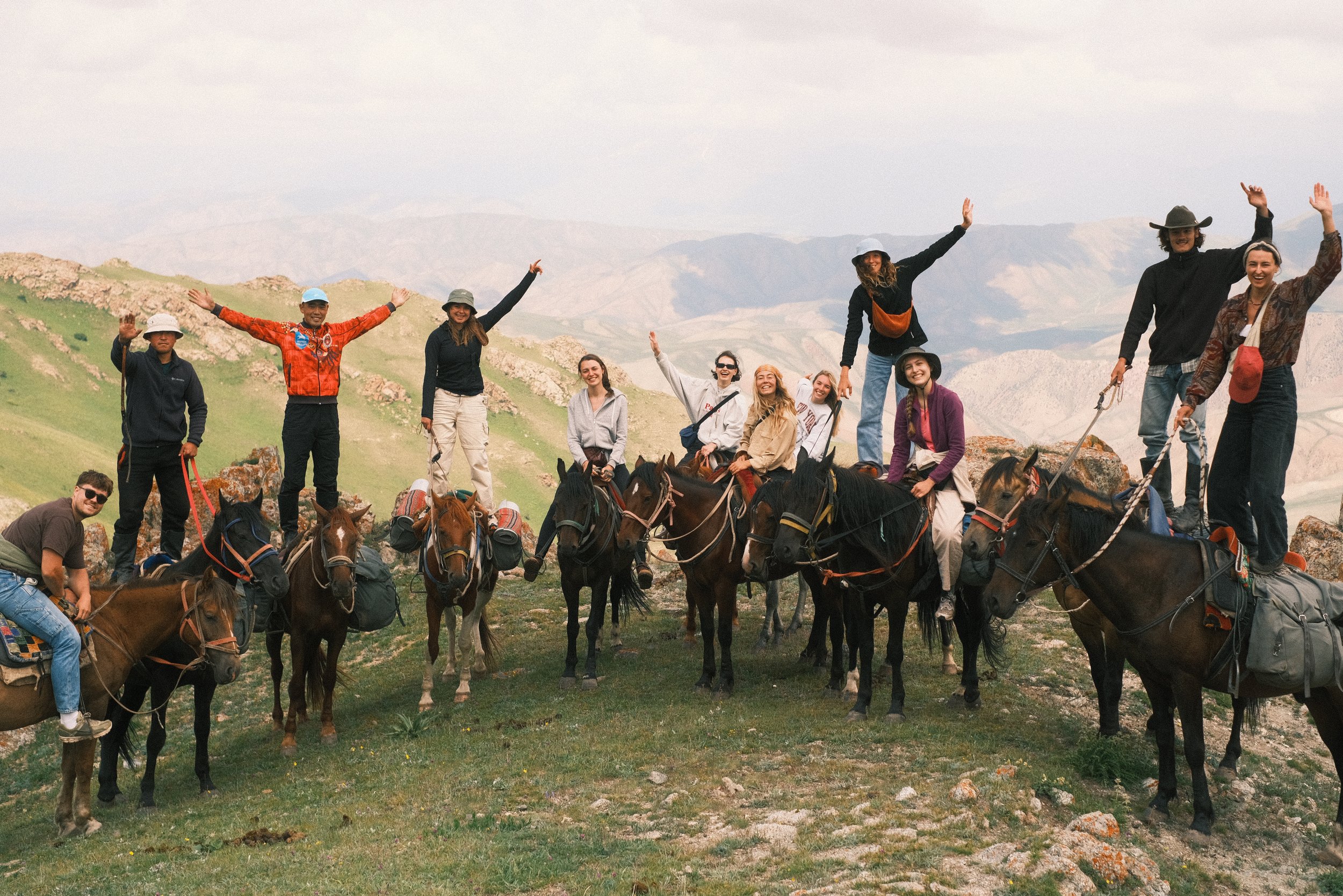 Group of people on horseback in a mountainous landscape, some standing on horses with arms raised and smiling, others sitting on horses, wearing outdoor clothing and hats.