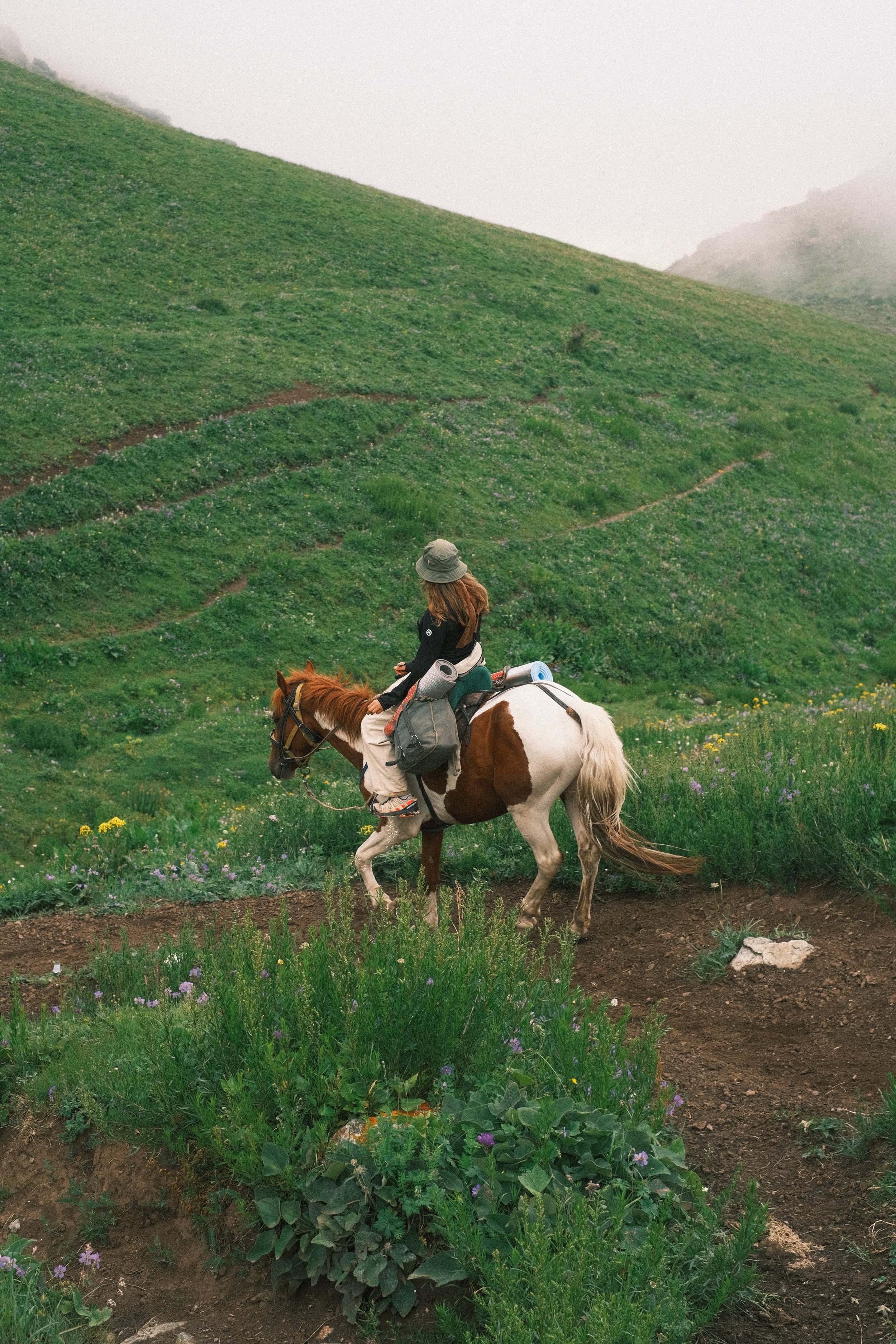 A person riding a white and brown horse on a trail through a green, hilly landscape with patches of wildflowers and fog covering the hilltops.