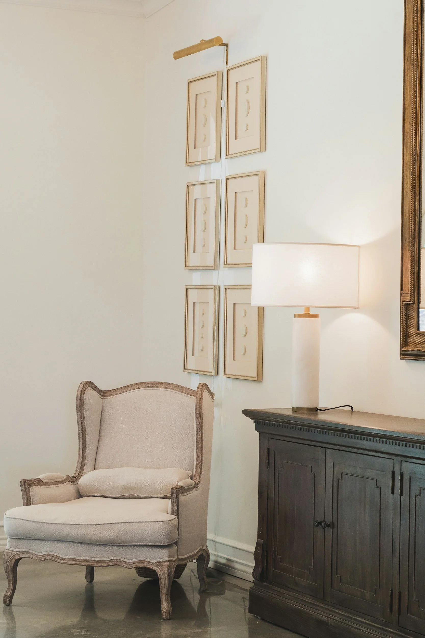 Cream-colored upholstered armchair with wooden trim beside a dark wood sideboard, white table lamp, and wall art with framed photos of moon phases.