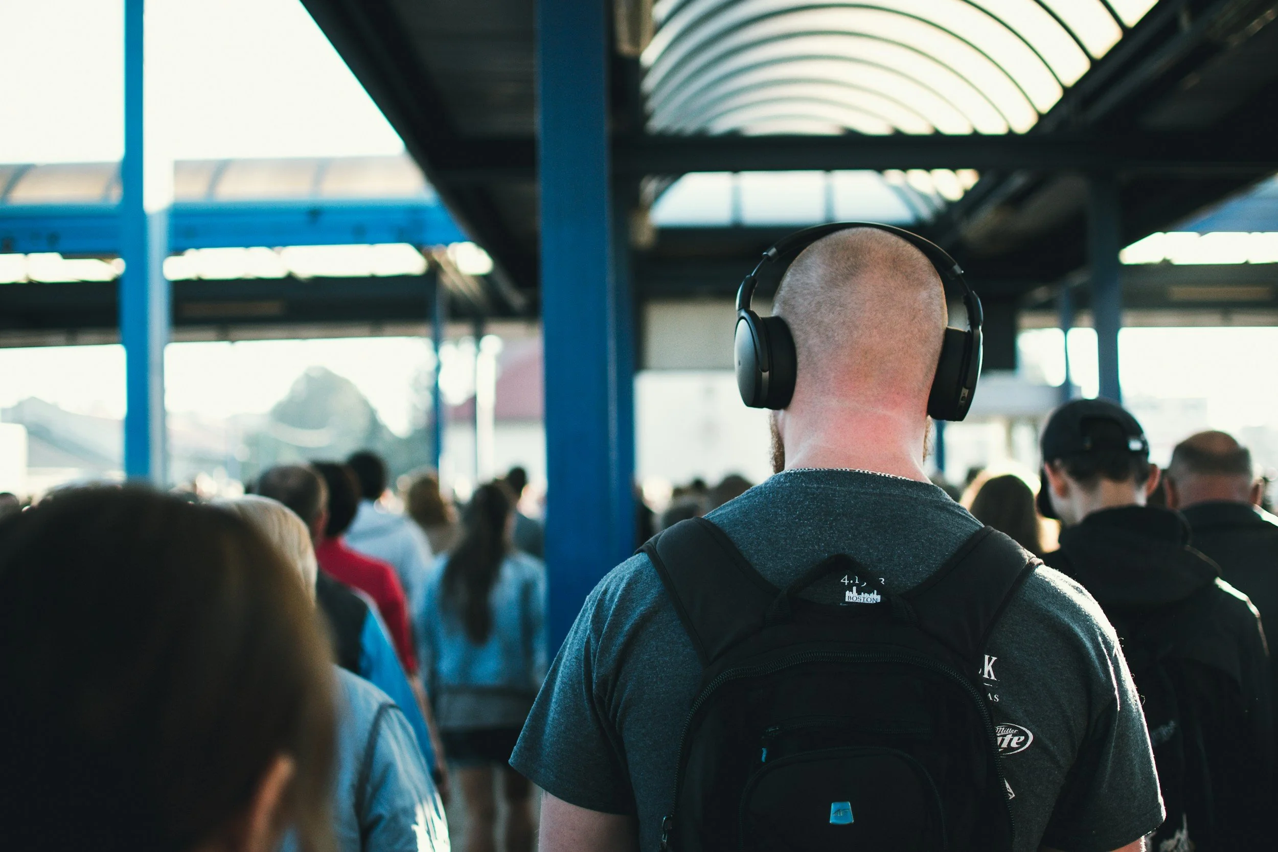 Back view of a man with a buzz cut and headphones, waiting in line at a crowded transportation station.