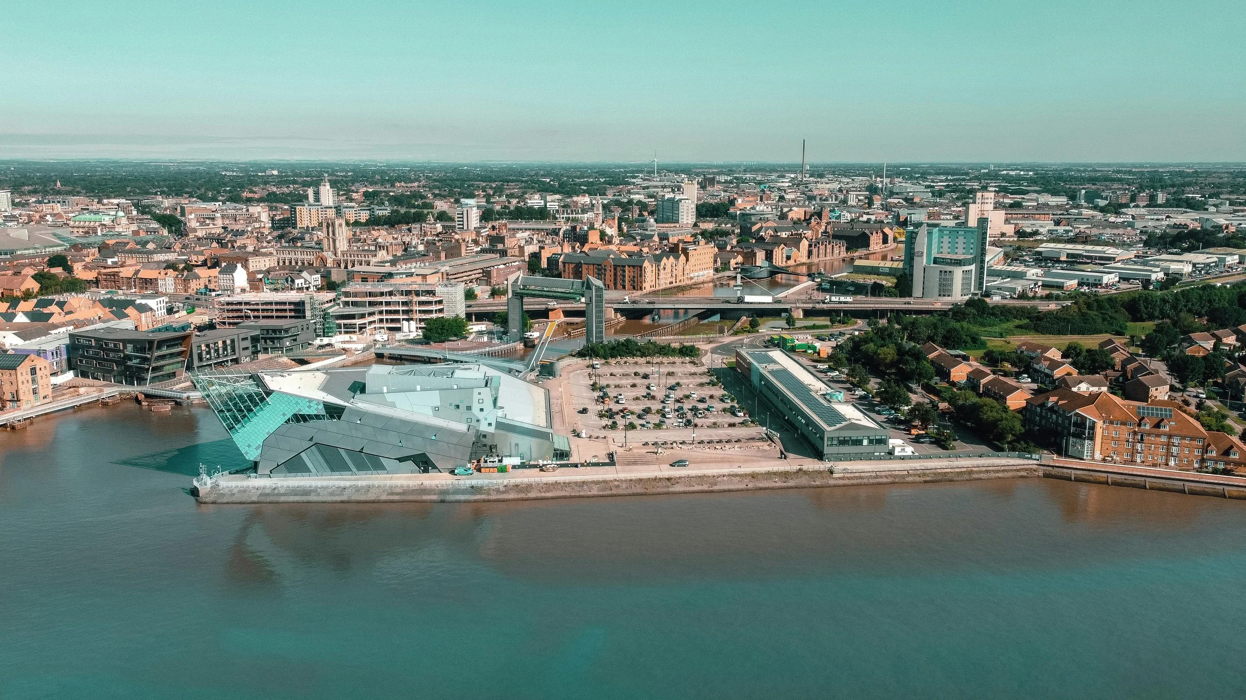 Aerial view of a city with modern buildings, bridges, a waterfront, and parking lots, featuring a distinctive angular glass building near the water.