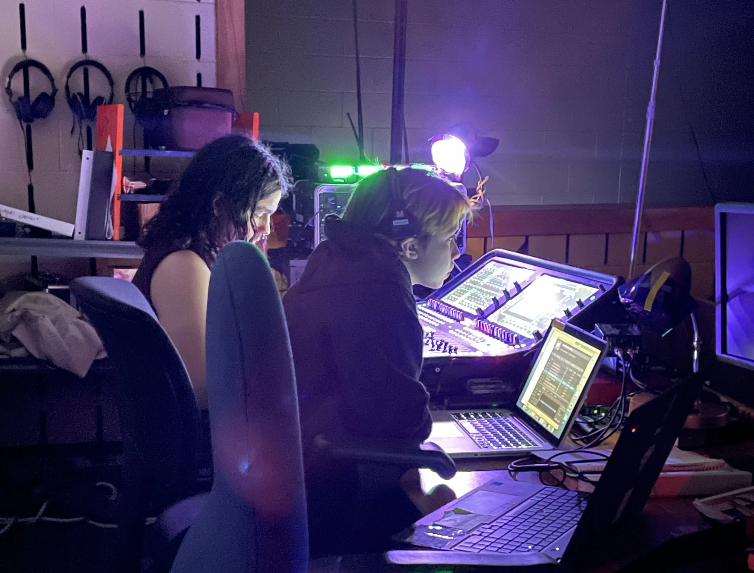 Two women are working at a mixing console and computers in a dimly lit room, possibly a recording or radio studio.
