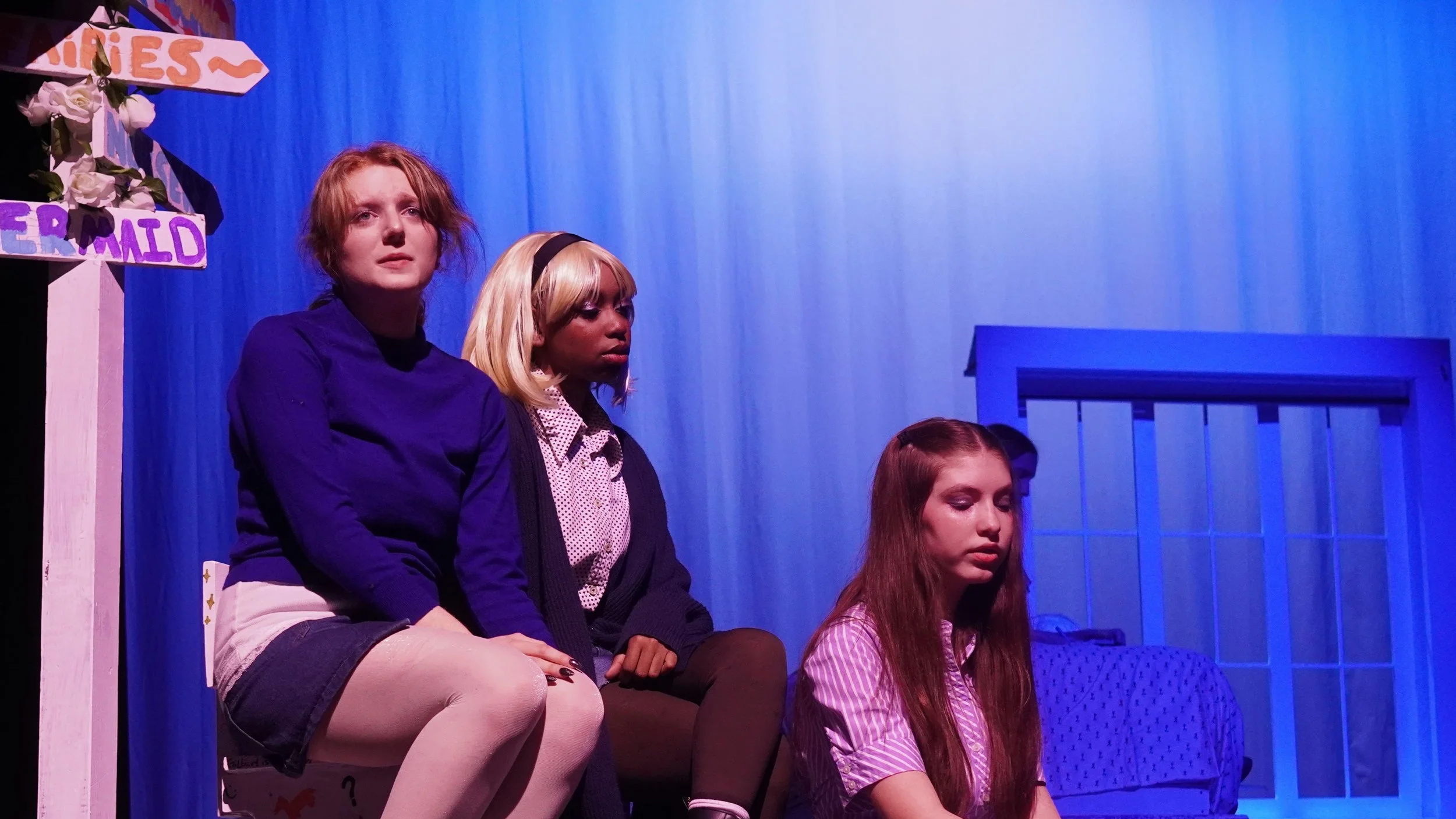 Three young women sitting on stage with blue backdrop, appearing somber or contemplative. One has short red hair, another has blonde hair with a headband, and the third has long brown hair.
