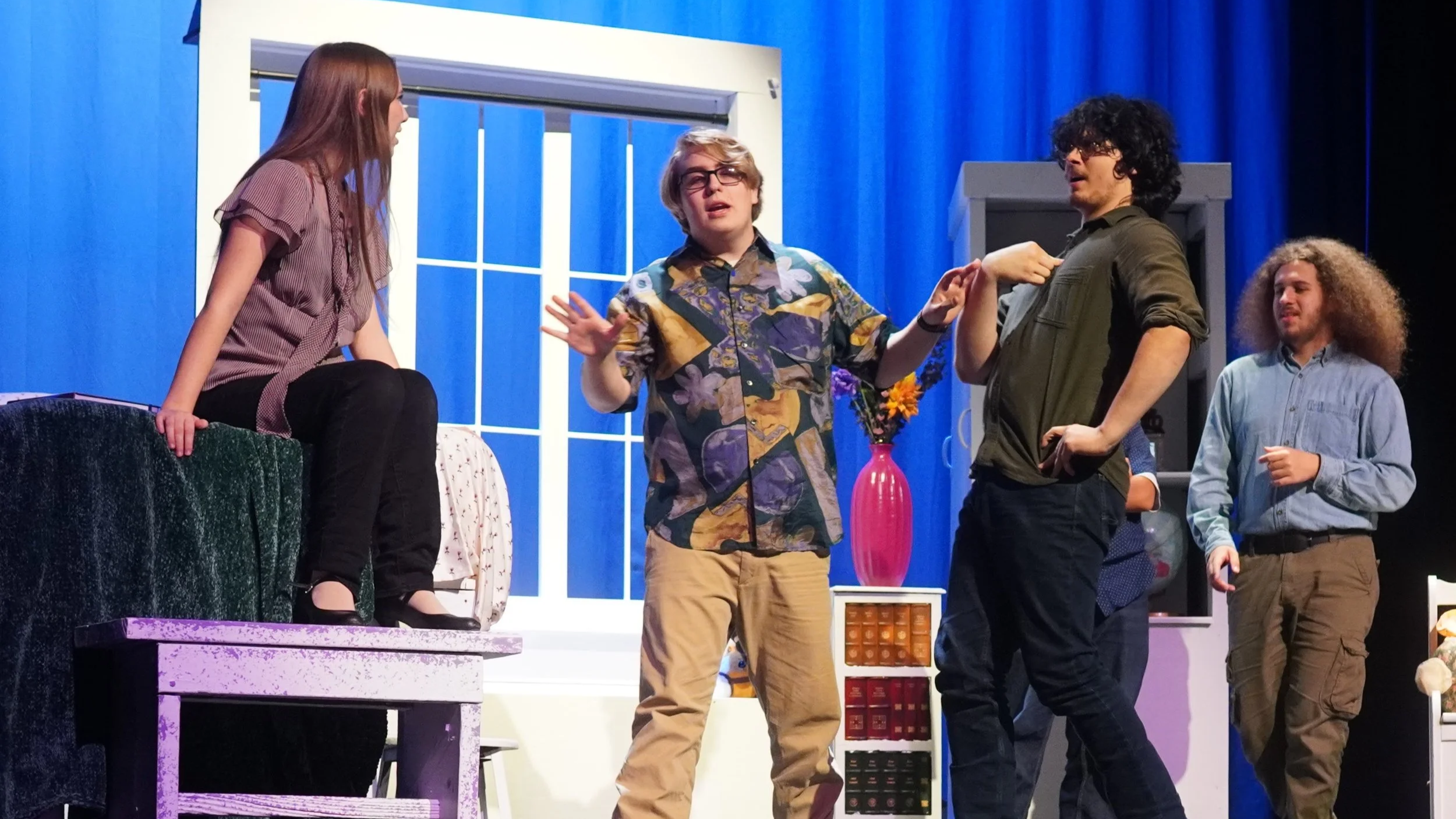 Four young actors perform in a scene on stage, with a woman sitting on a green-colored prop, a blue curtains backdrop, and a window set piece. One actor stands in the center gesturing with hands, while the others watch.