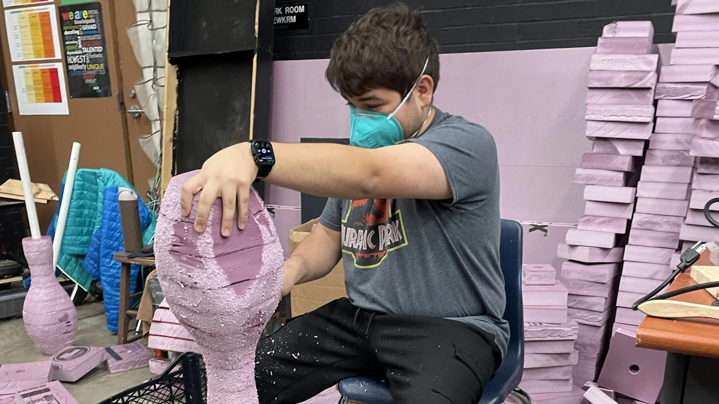 A young man wearing a green face mask and a smartwatch is sculpting a large, pink foam sculpture in a workshop. The workshop is filled with stacks of foam and sculptures, and there are craft tools visible on a nearby table.