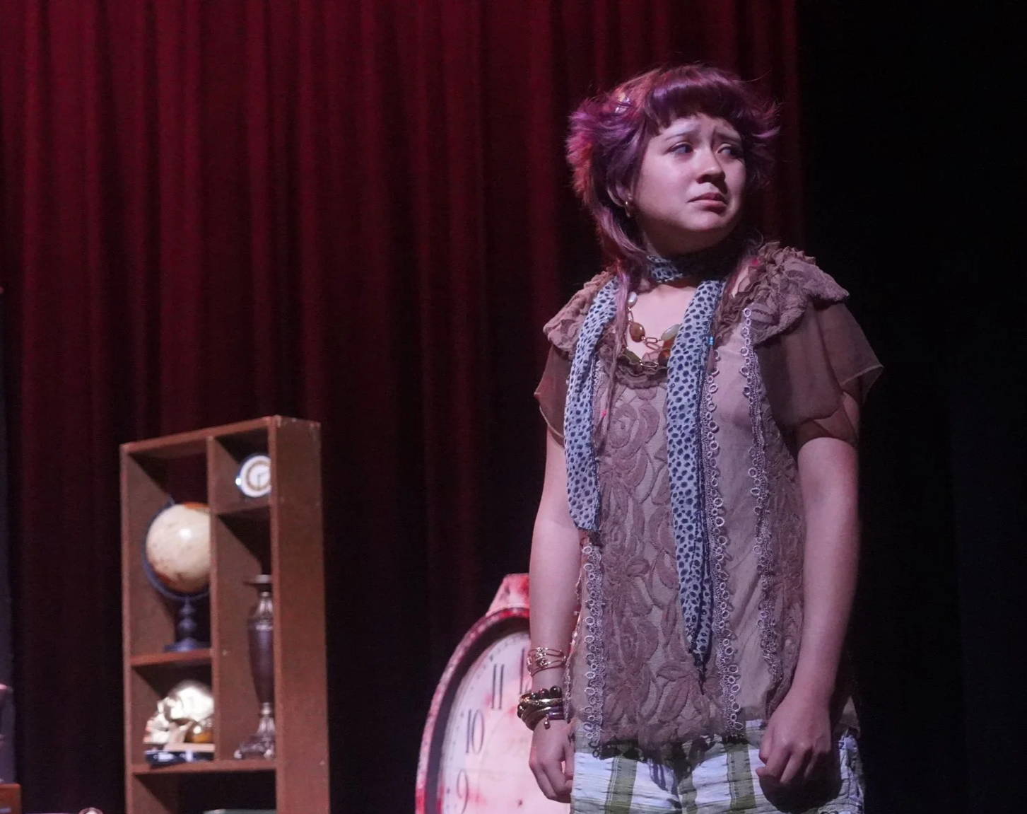 Young woman performing on stage with a red curtain in the background, a clock, and a shelf with decorative items.