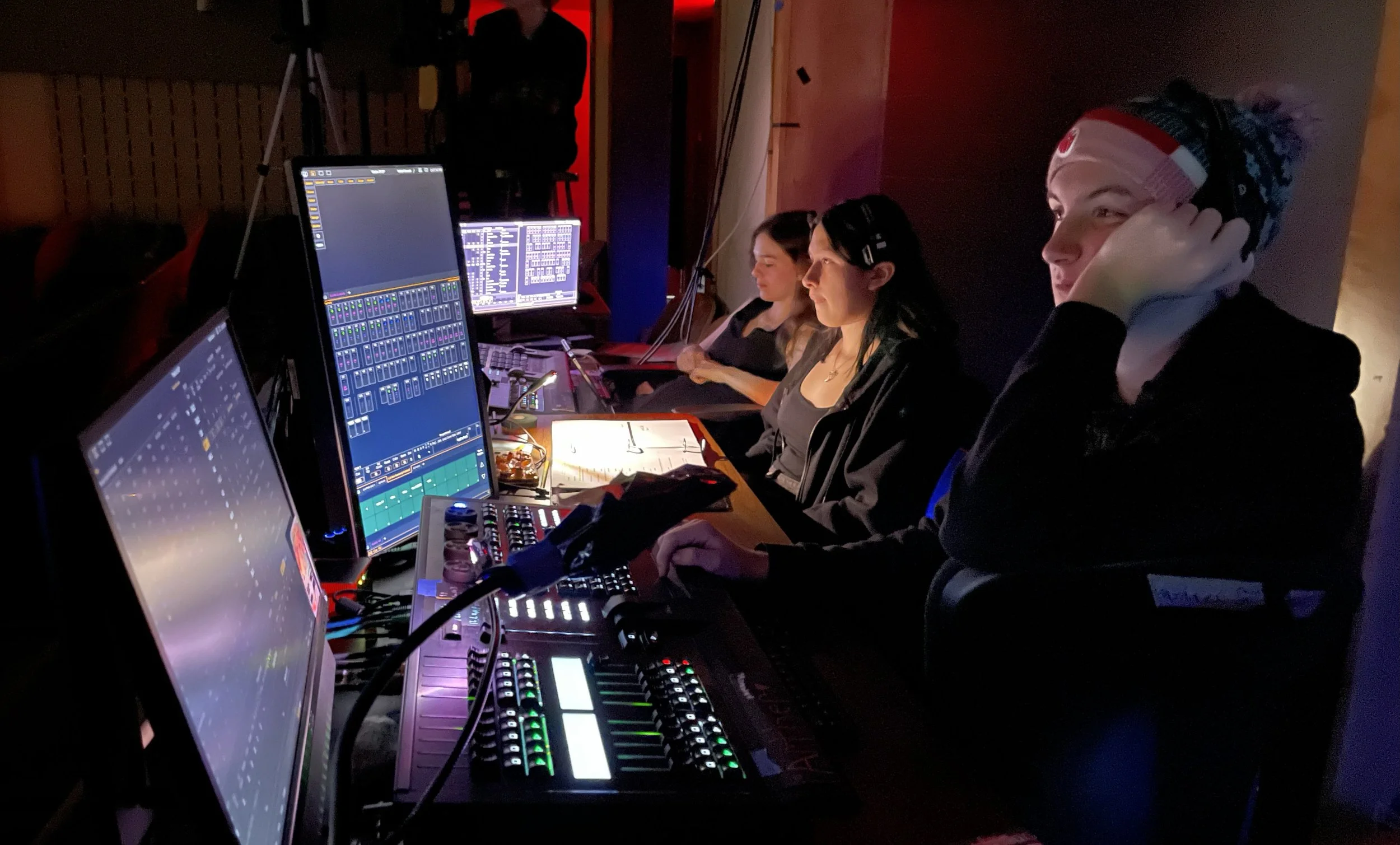 Four people working at a audio mixing control board with multiple monitors in a dark room, with some wearing headphones.