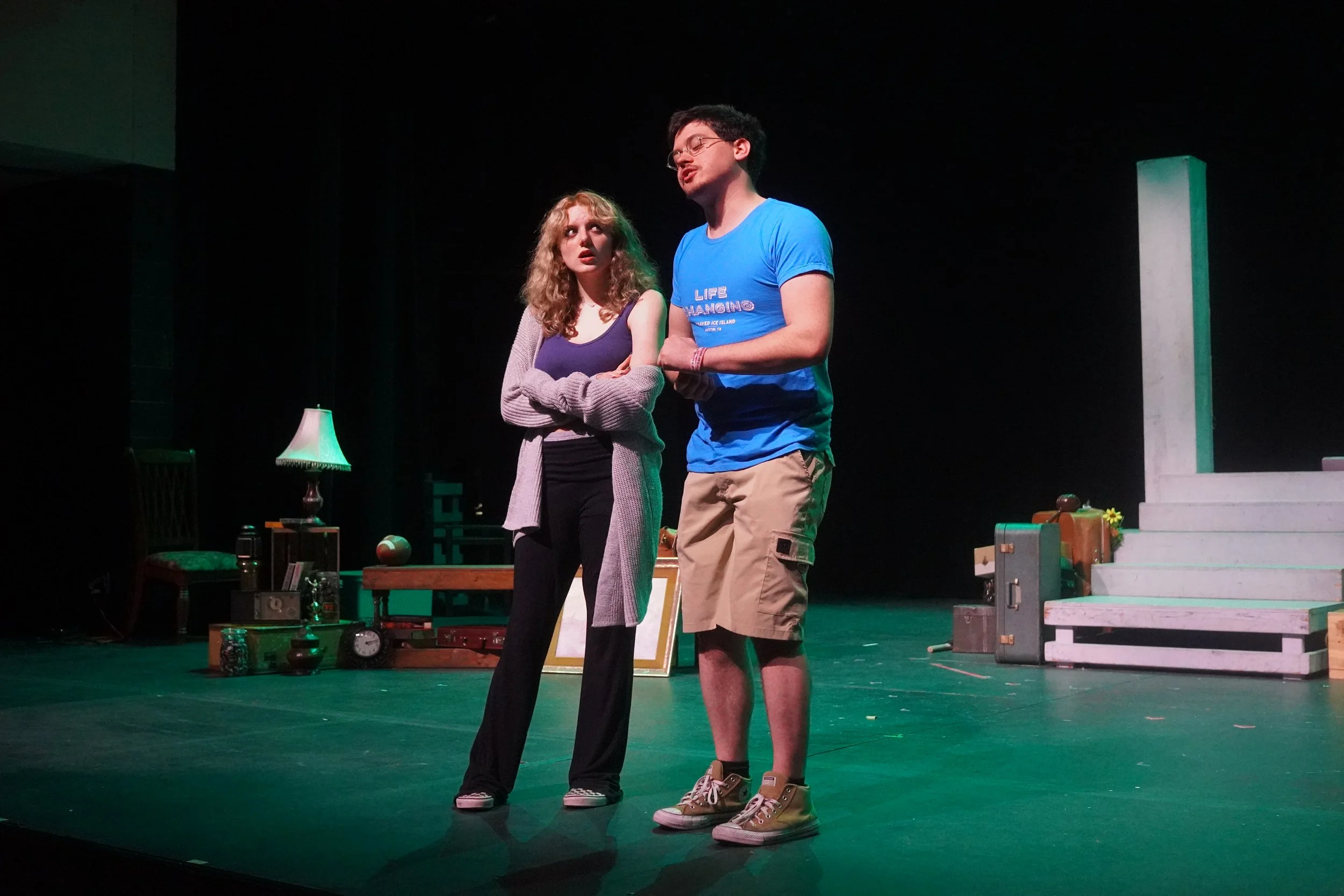 Two actors perform on stage; a woman with curly blonde hair, wearing a purple top and black pants, stands with arms crossed looking concerned, and a man with dark hair and glasses, wearing a blue t-shirt and beige shorts, is speaking with eyes closed