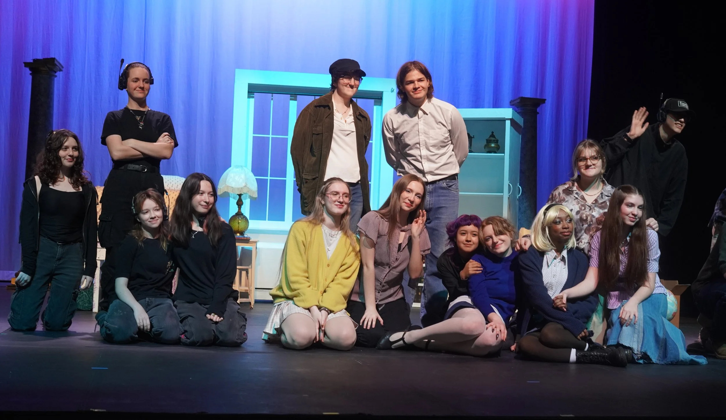 Group of young people on stage in a theatrical production, some standing and some sitting, with colorful background and set pieces.
