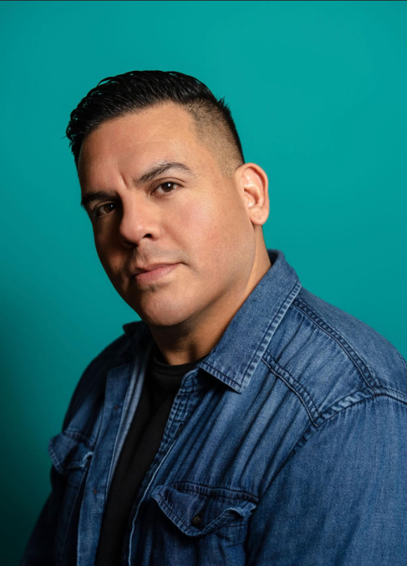 A portrait of a man with short dark hair, wearing a denim jacket over a black shirt, against a teal background.
