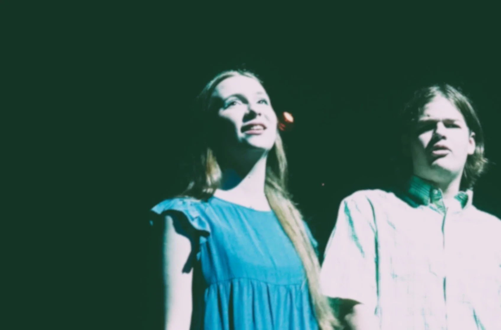 Two young people, a woman and a man, looking upward with expressions of wonder or surprise, standing side by side against a dark background.