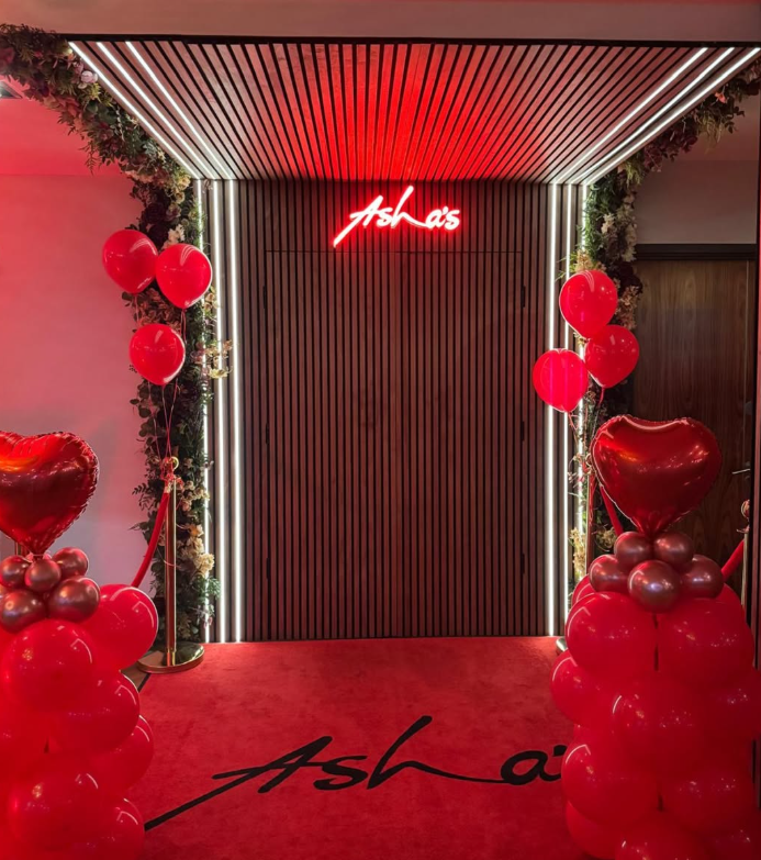 Decorated entrance with red and heart-shaped balloons, floral garlands, and a neon sign that reads 'Asha's' in red with a red carpet.