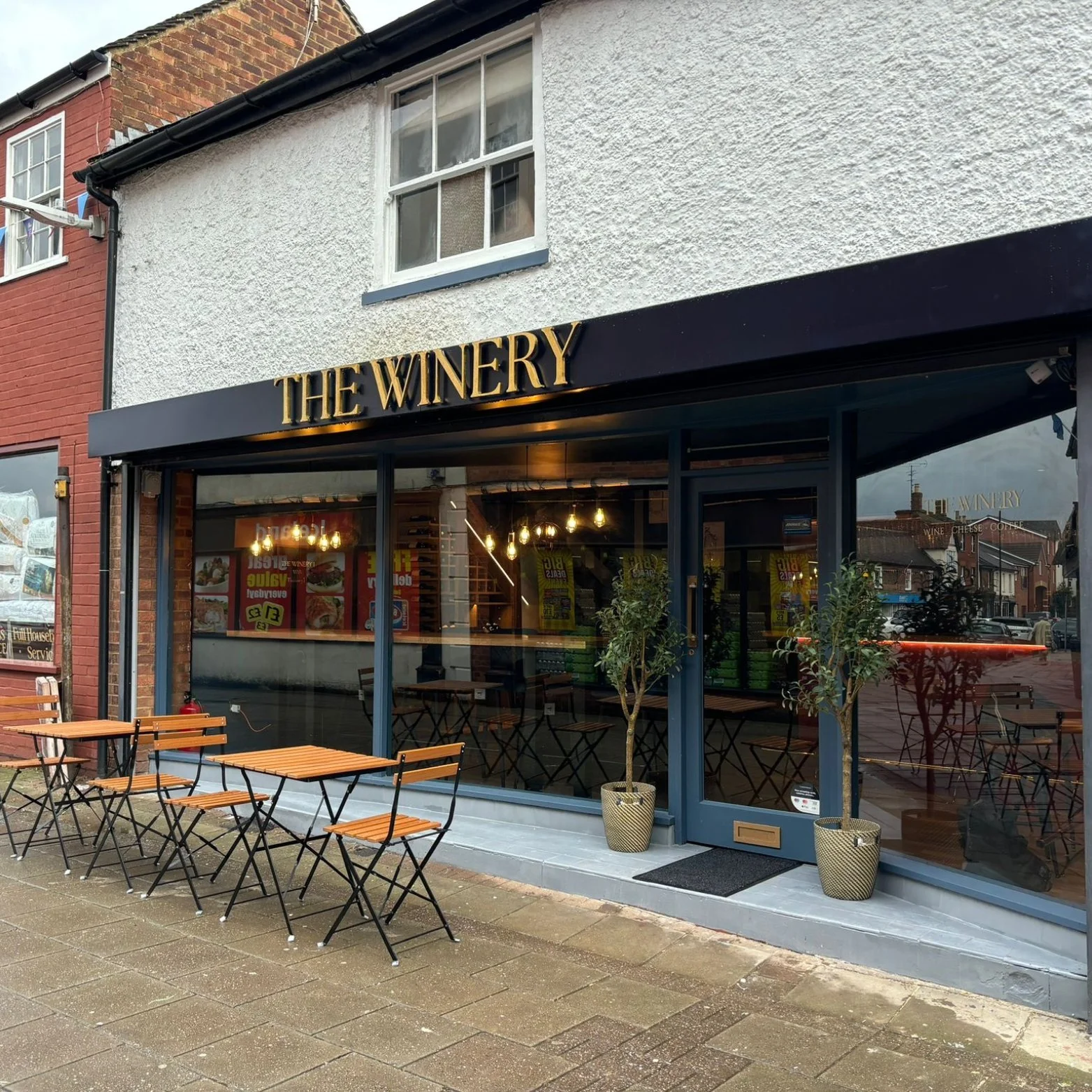 Exterior view of a restaurant called 'The Winery' with outdoor seating, including three small tables with matching chairs, and two potted trees in front of large glass windows.