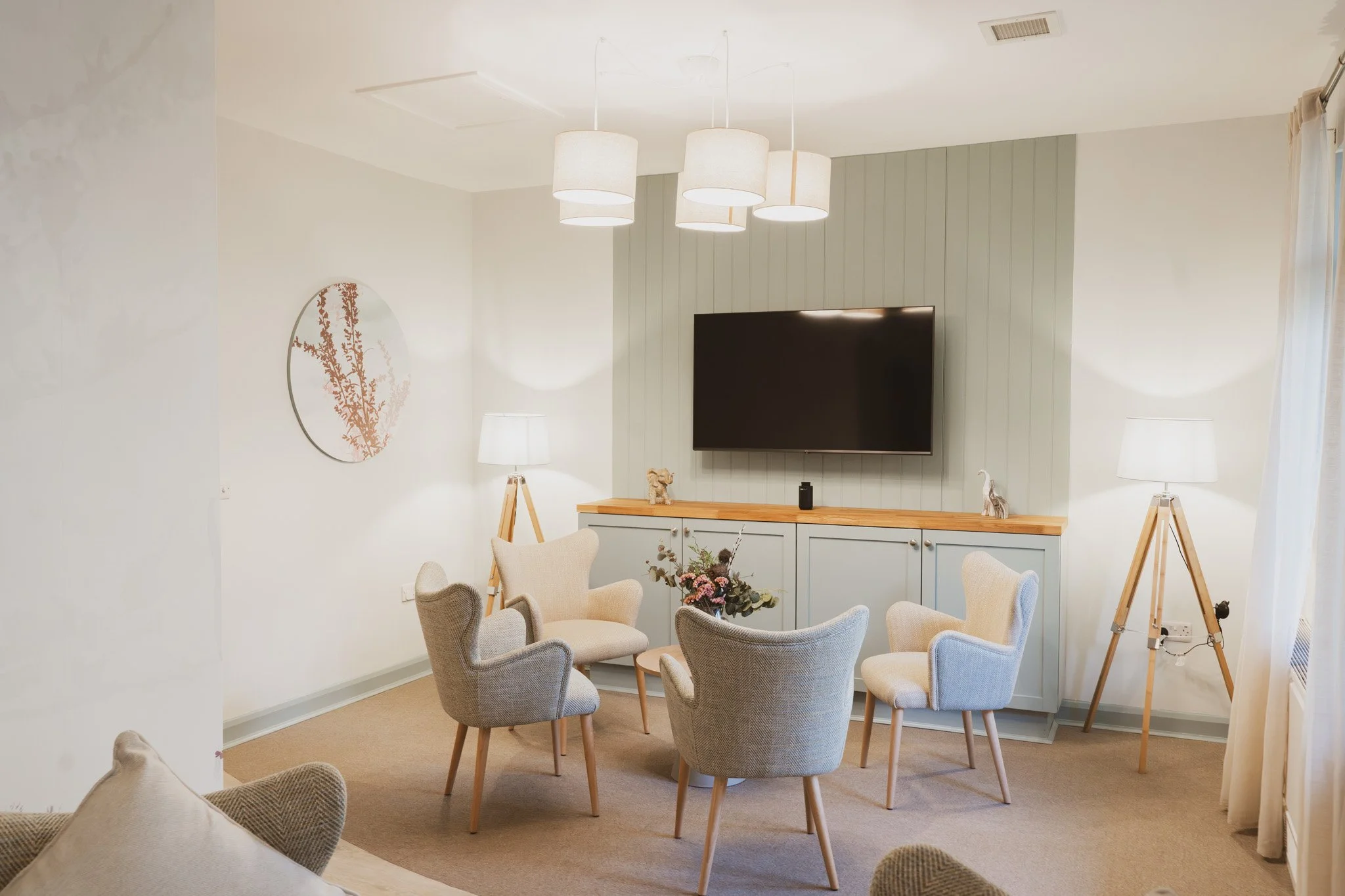 Living room with six beige and gray upholstered armchairs arranged in a circle around a floral centerpiece on a small wooden table. A large flat-screen TV is mounted on the wall above a cabinet with decorative items, flanked by two floor lamps. Ceiling has white and beige drum pendant lights, and a window with light-colored curtains is visible on the right.