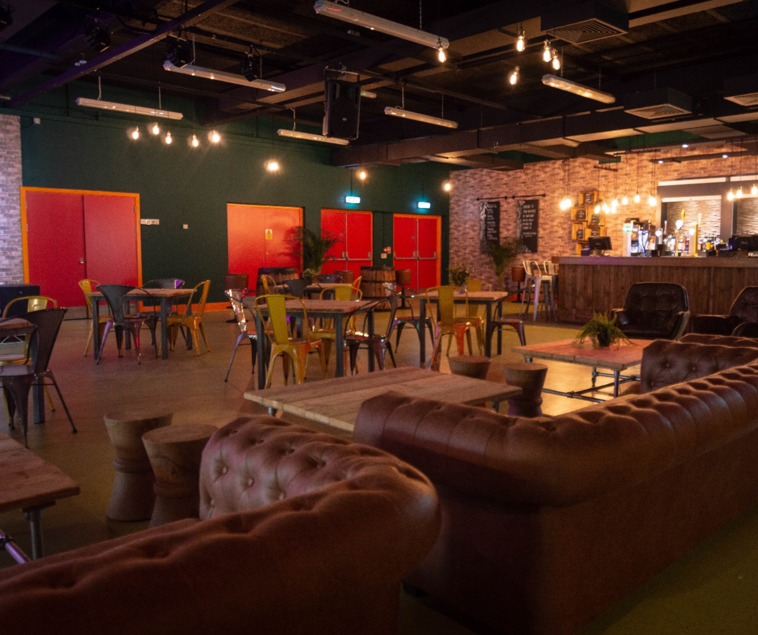 Interior of a dimly lit bar or lounge with wooden furniture, leather sofas, colorful metal chairs, a bar counter, and exposed brick walls, illuminated by hanging light bulbs.