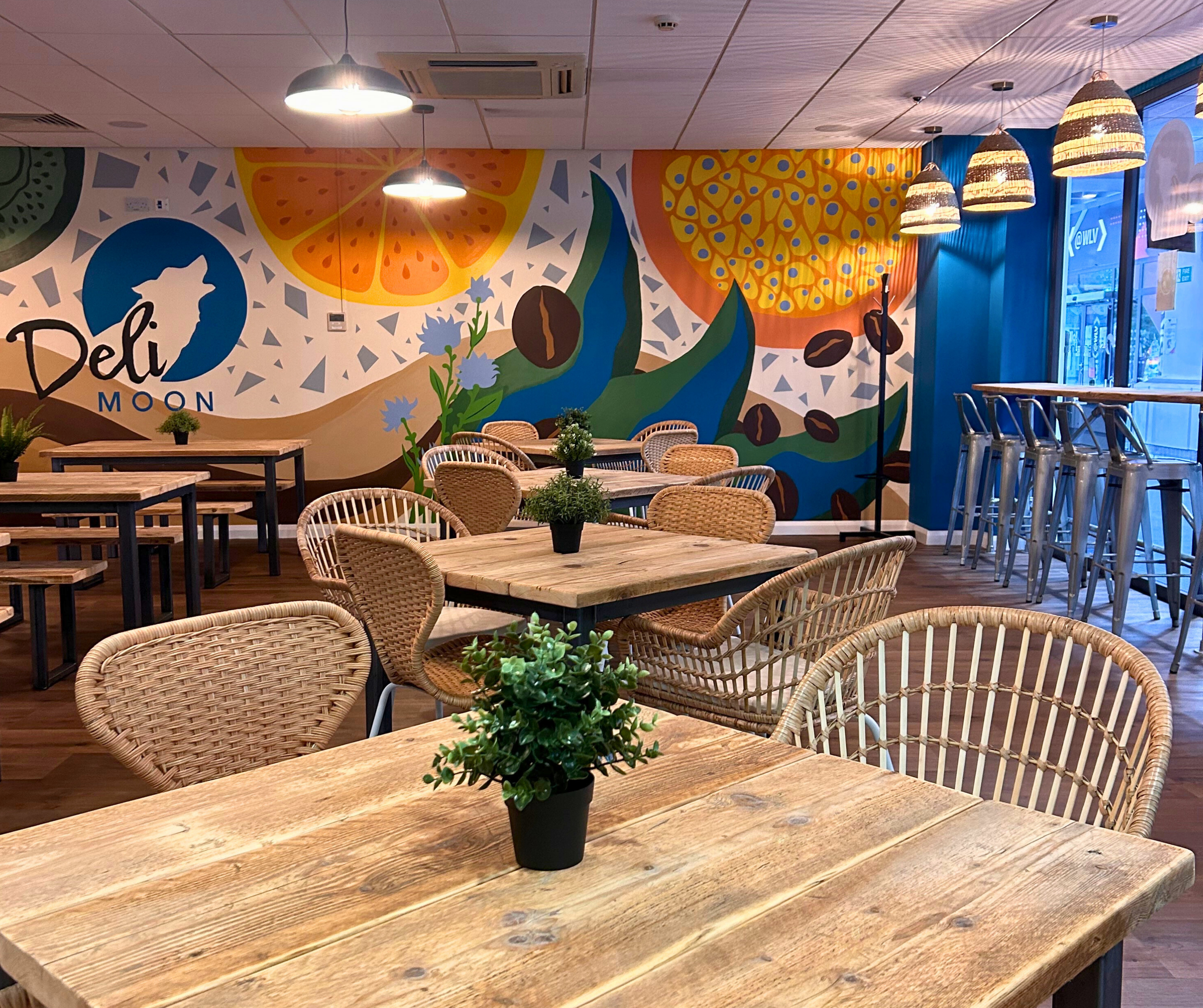 Interior of a cafe or restaurant with rattan chairs and wooden tables decorated with small potted plants, colorful mural on the wall featuring fruit and coffee beans, large windows with blue curtains, and hanging lights.