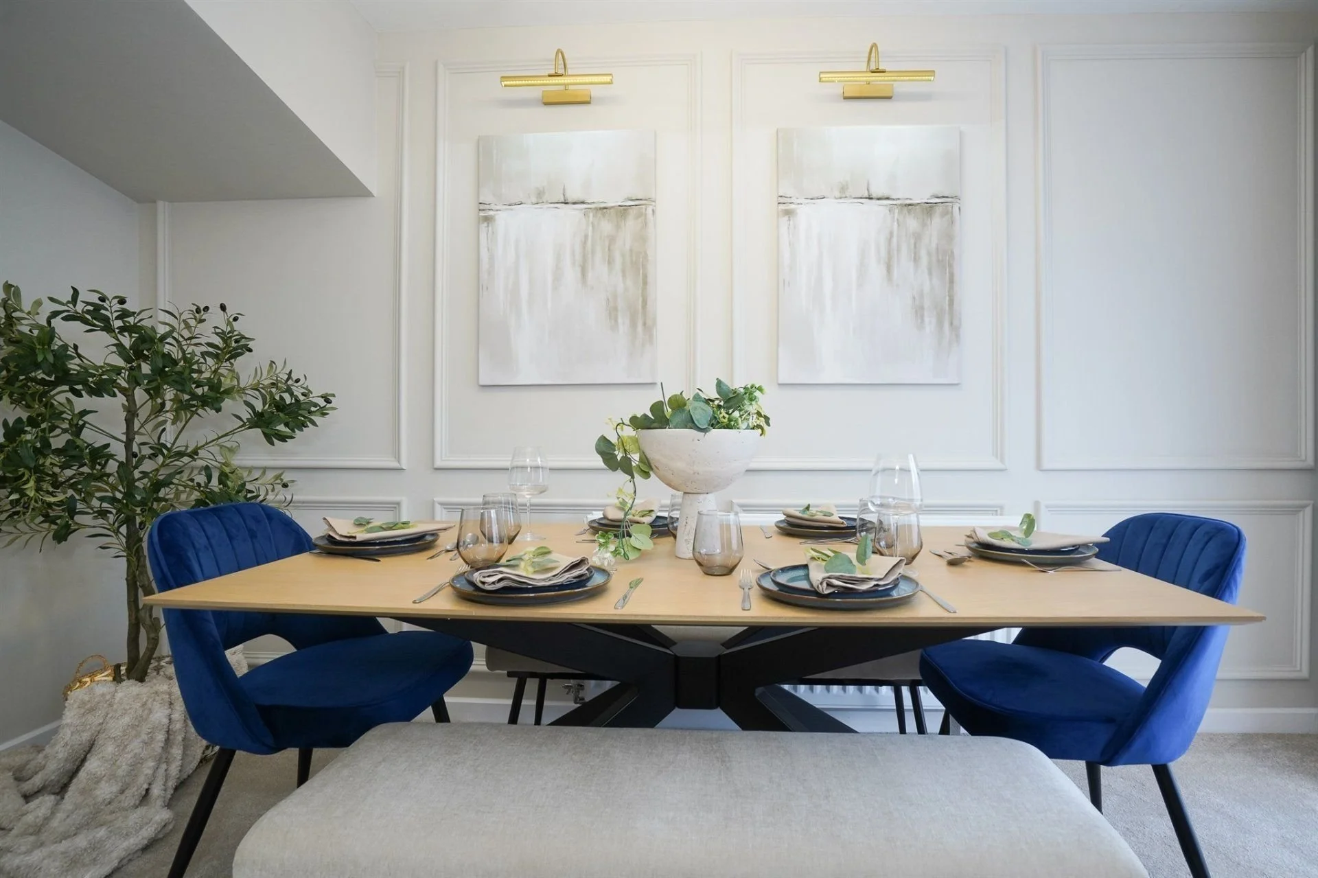 Dining room with a wooden table set for six, blue velvet chairs, a large white vase with greenery, modern art paintings, and gold wall sconces.