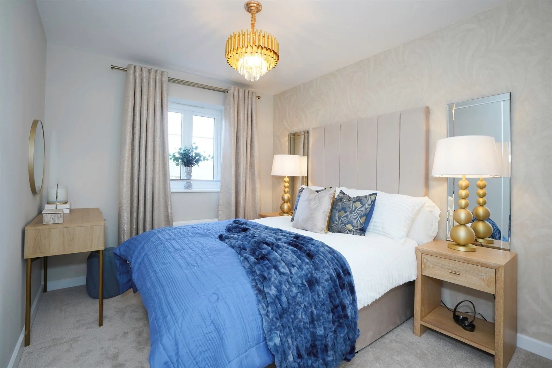 A bedroom with a bed dressed in white and blue bedding, four throw pillows, a beige headboard, side tables with gold lamps, a window with beige curtains, a chandelier, a mirror, and a small desk with decorations.