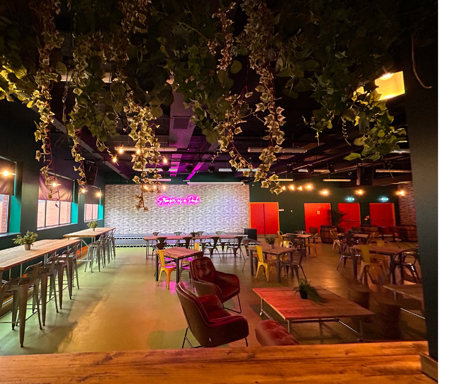 An empty lounge or event space decorated with hanging greenery, neon pink sign, various chairs and tables, with a brick wall and red doors in the background.