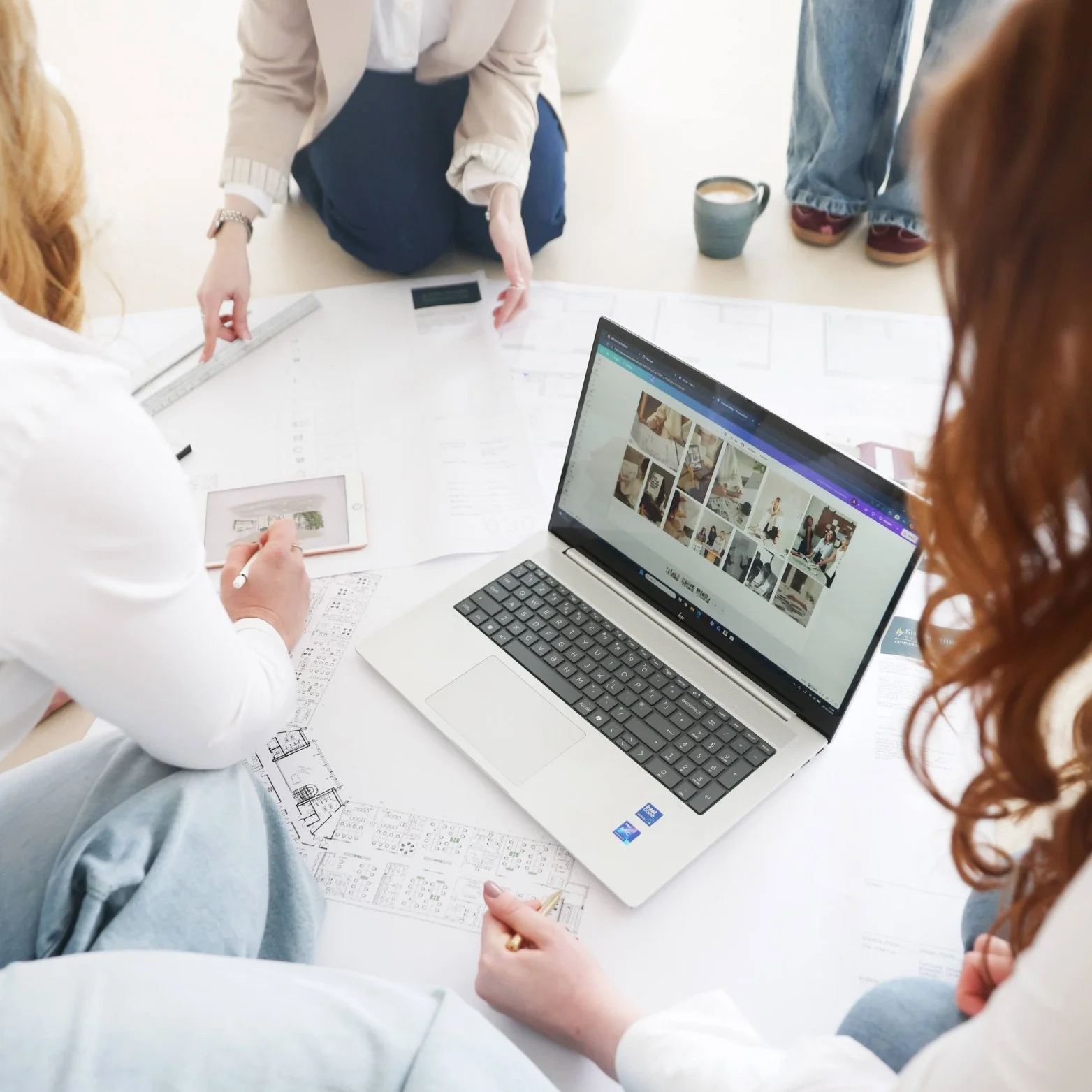 Group of people working on a project with architectural plans, a laptop displaying photos, a tablet, and a mug on a table.