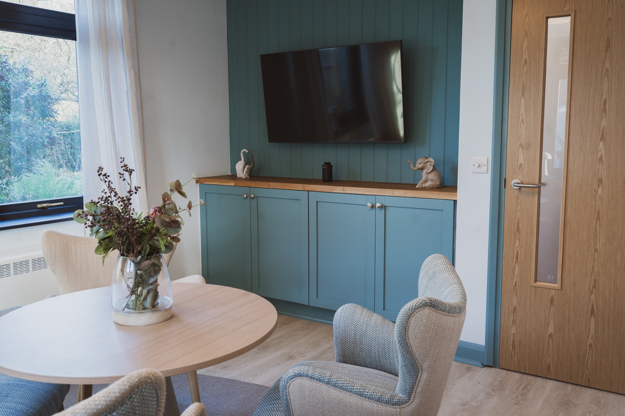 Living room with a round wooden table, a vase with flowers, and beige upholstered chairs, with a wall-mounted TV, decorative elephant statues, a blue cabinet with a wooden top, a window with white curtains, and a wooden door.
