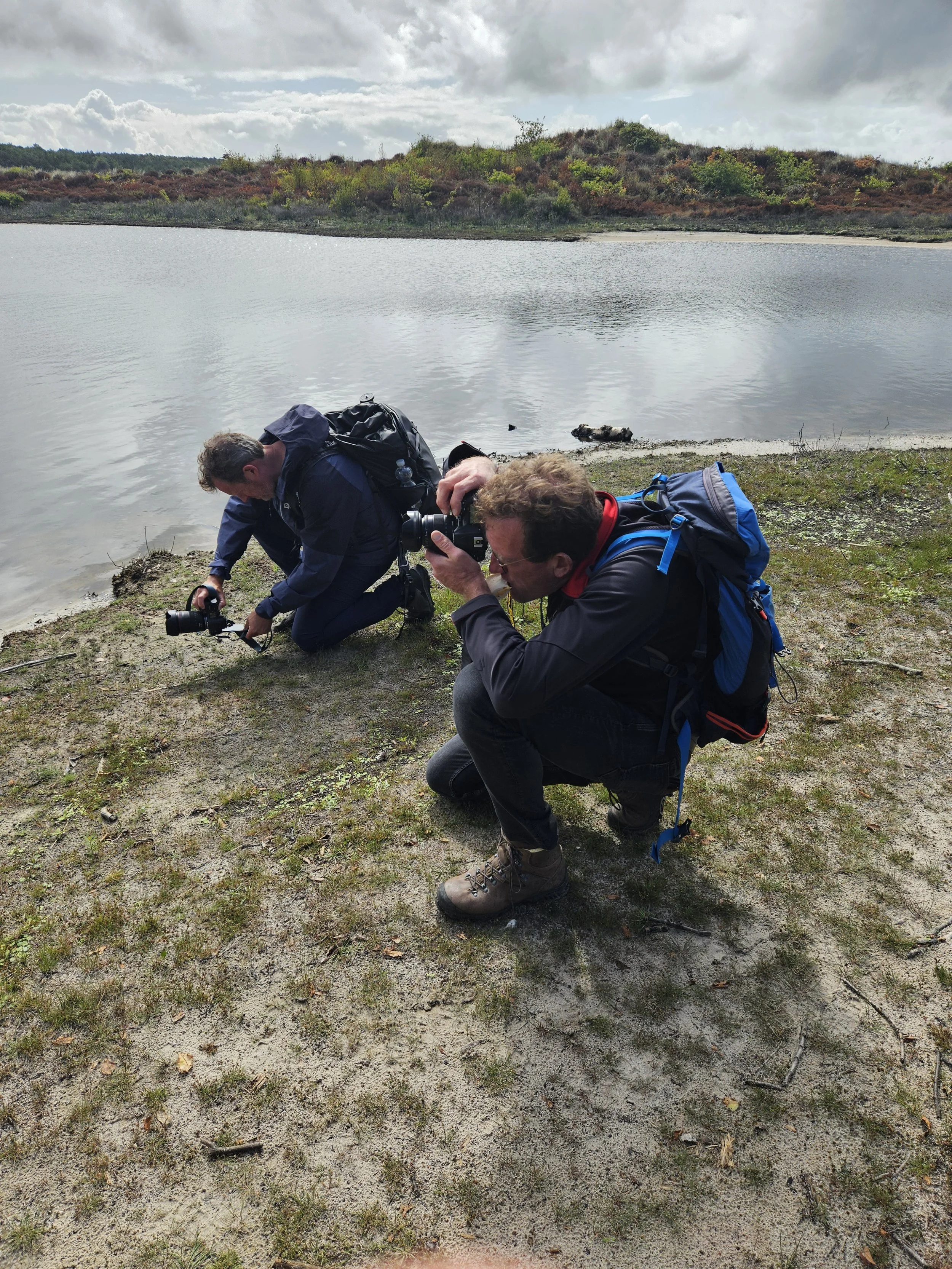 Twee fotografen buigen zich laag bij laag aan de rand van een meer, bezig met fotograferen, in een natuurlijke omgeving met water, zand en struiken.
