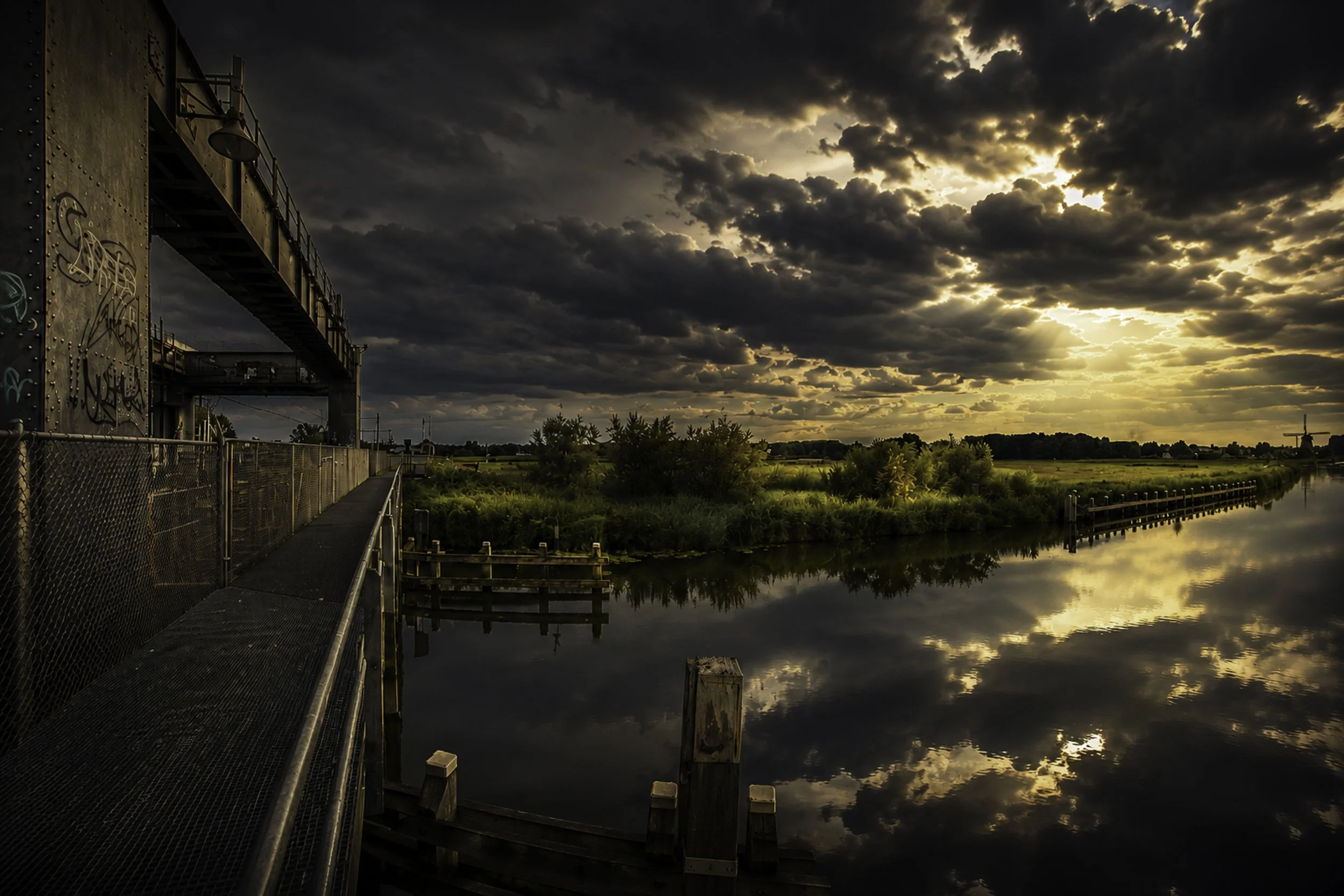 Een haven of kade met een brug en water, onder een donkere, bewolkte hemel met de zon die door de wolken schijnt.