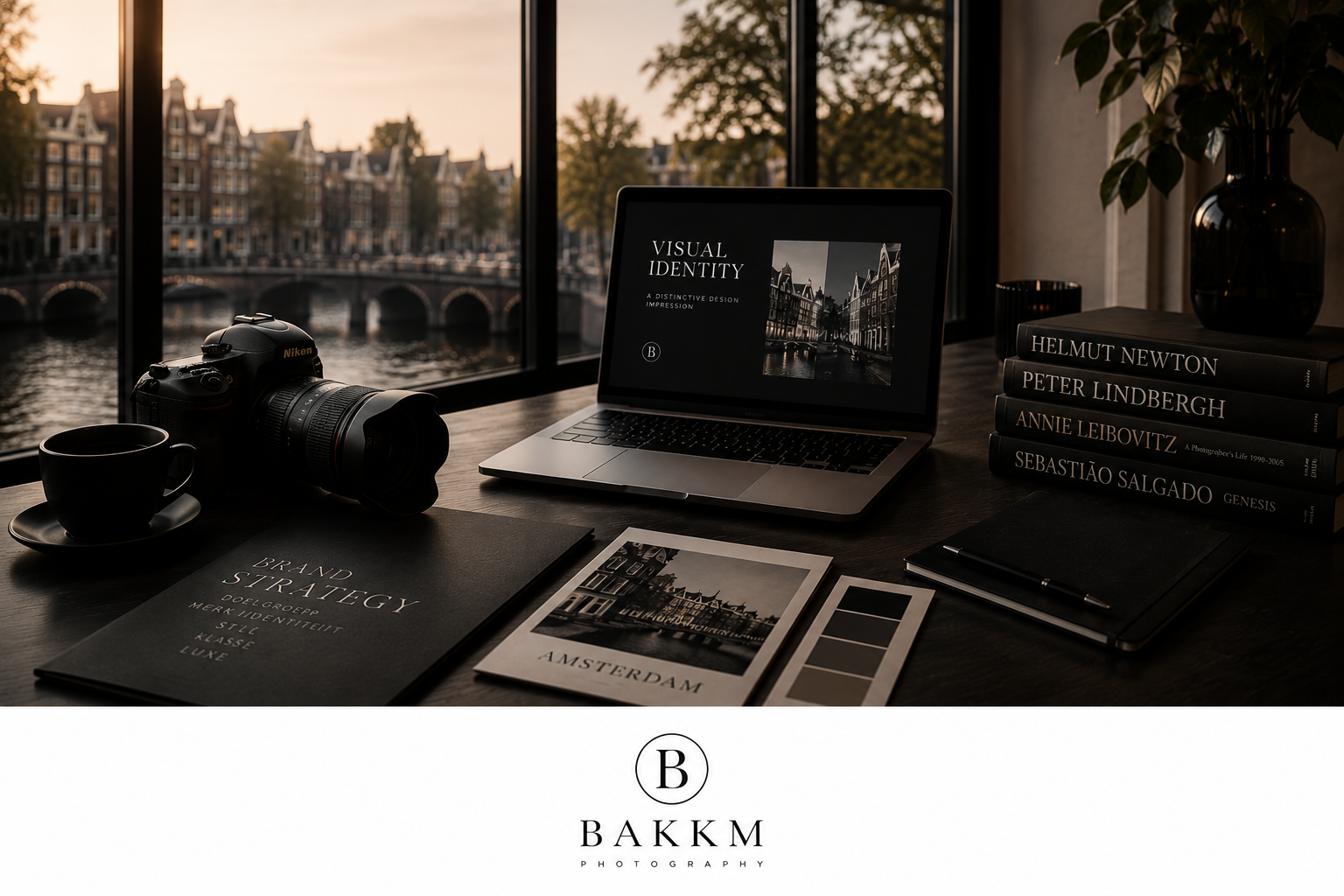 A workspace with a Nikon camera, a laptop displaying a cityscape with the words "Visual Identity," a black coffee cup on a saucer, a portfolio titled "Brand Strategy," printed photographs, a design book, a closed notebook with a pen on top, and a stack of art books seen through large windows overlooking a canal with historic buildings.