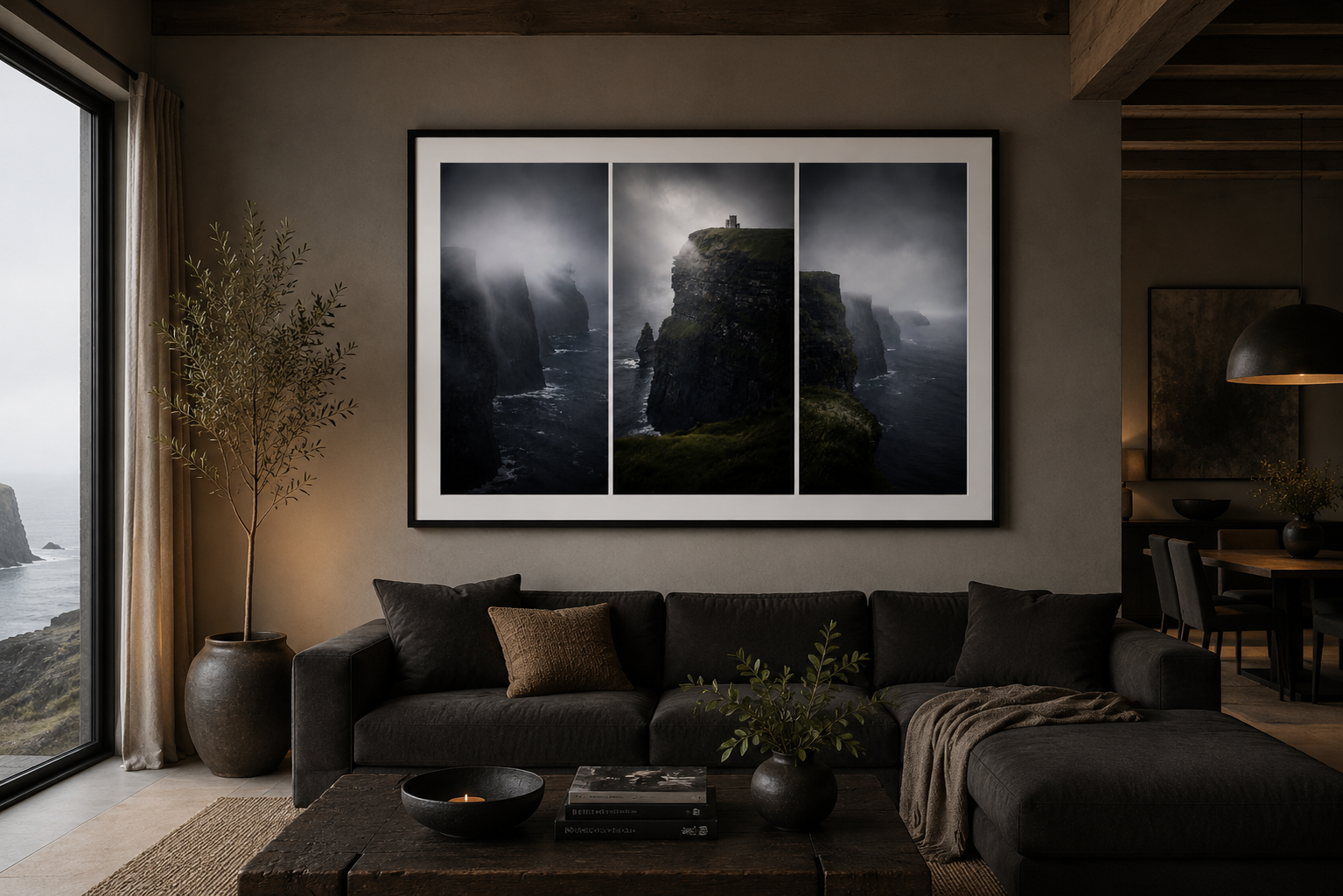 Living room with black sofa, large window showing ocean view, large framed triptych photo of rugged coastal cliffs, potted plant, dark coffee table with books and a small candle, subdued lighting.