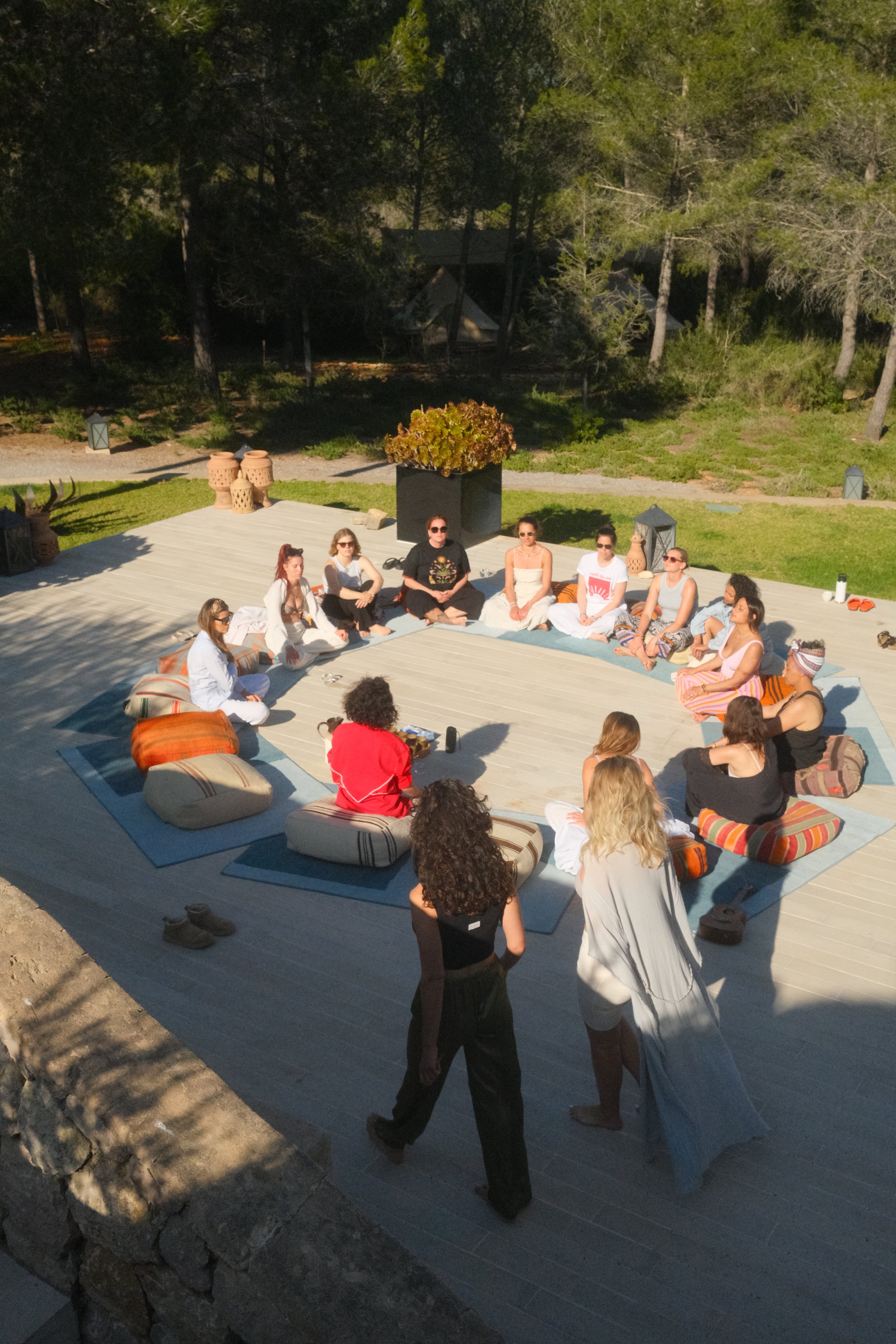 Menschen sitzen im Kreis auf einem Holzdeck im Freien, umgeben von Bäumen, es ist vermutlich Sonnenuntergang oder -aufgang.