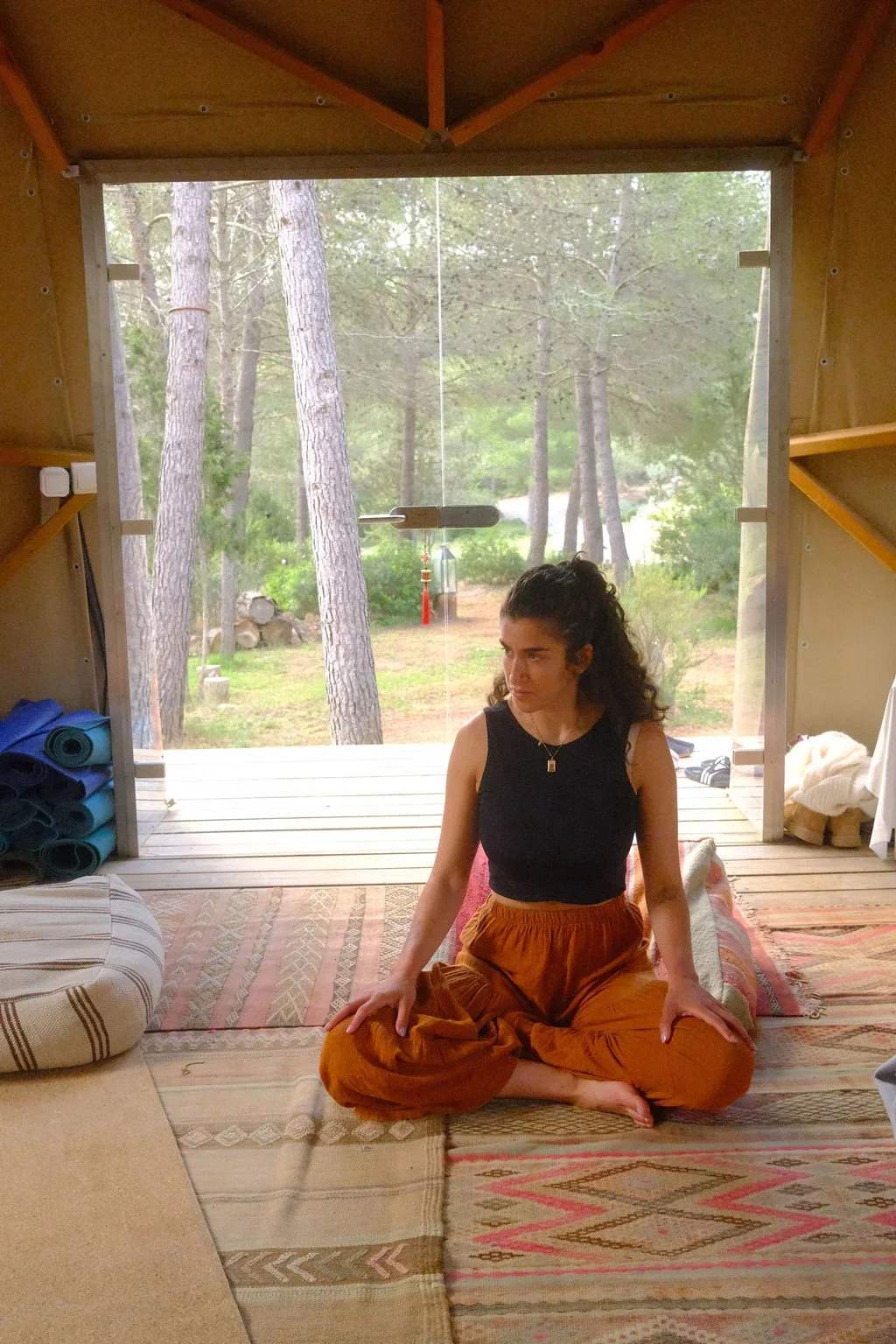 Eine junge Frau sitzt im Meditationsraum auf einem Teppich, mit Blick nach draußen auf einen Wald mit Bäumen.