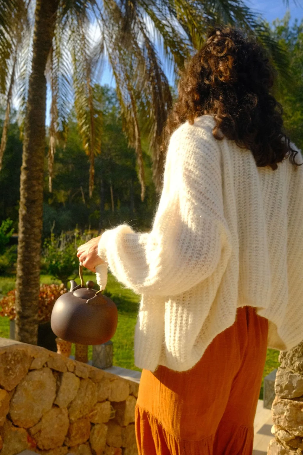 Frau in weißem Pullover und orangefarbenen Hosen hält eine dunkle Kaffeekanne im Garten mit Palmen im Hintergrund.